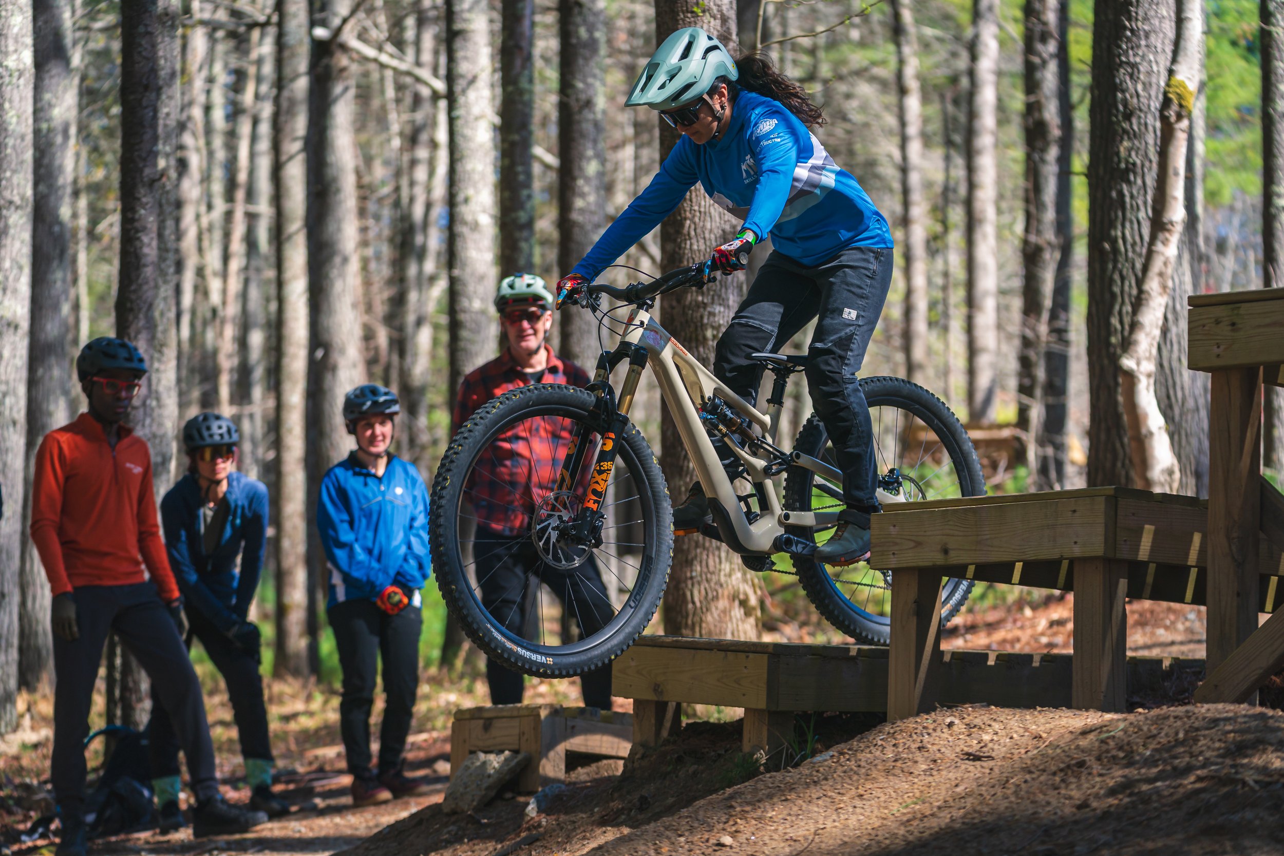 a mountain biker rides off a drop with her class watching from the side