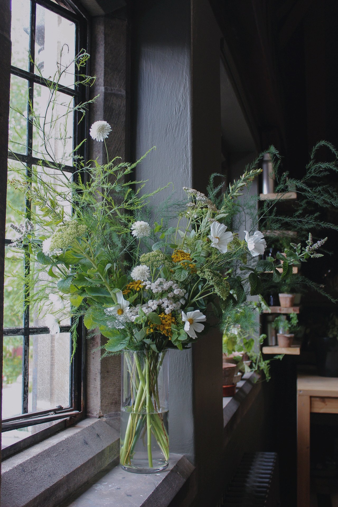 A glass vase filled with a mixed bouquet of white and green flowers is placed on a windowsill inside a room, for wedding venues. The window has black metal framing, and outside, trees and greenery are visible.