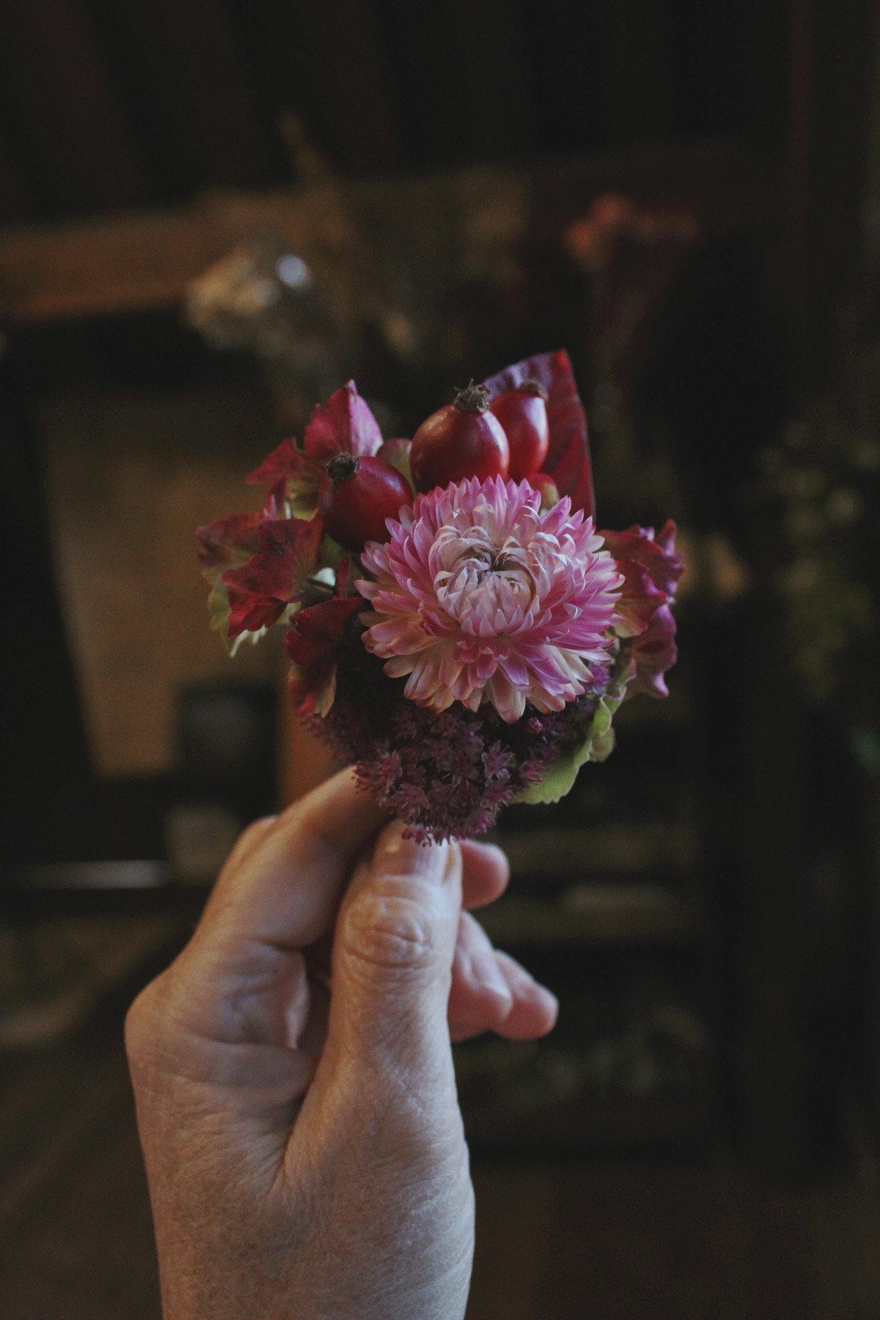 Hand holding a grooms buttonhole bouquet of pink and red flowers with berries, against a dark indoor background.