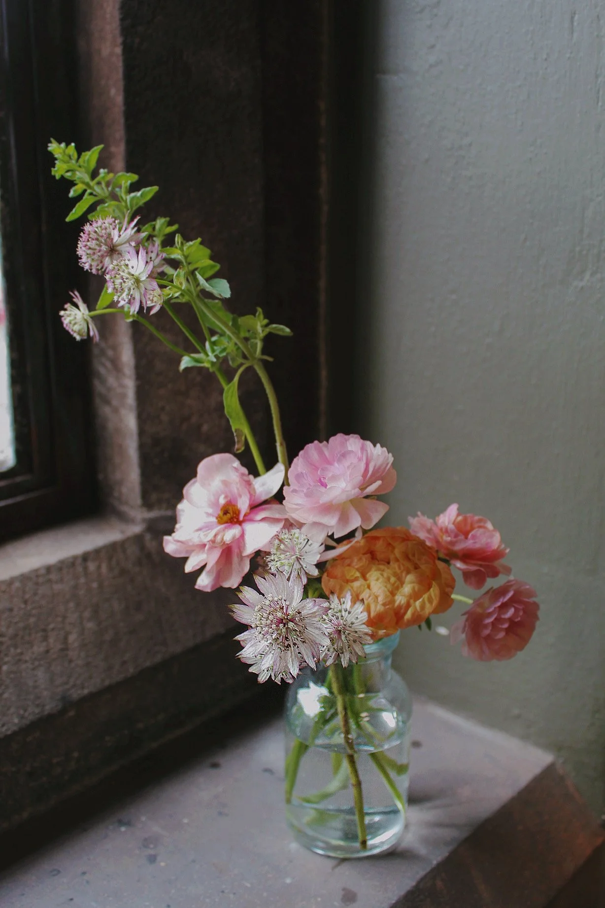 Small glass bud vase filled with delicate pink and white seasonal flowers on a stone windowsill.