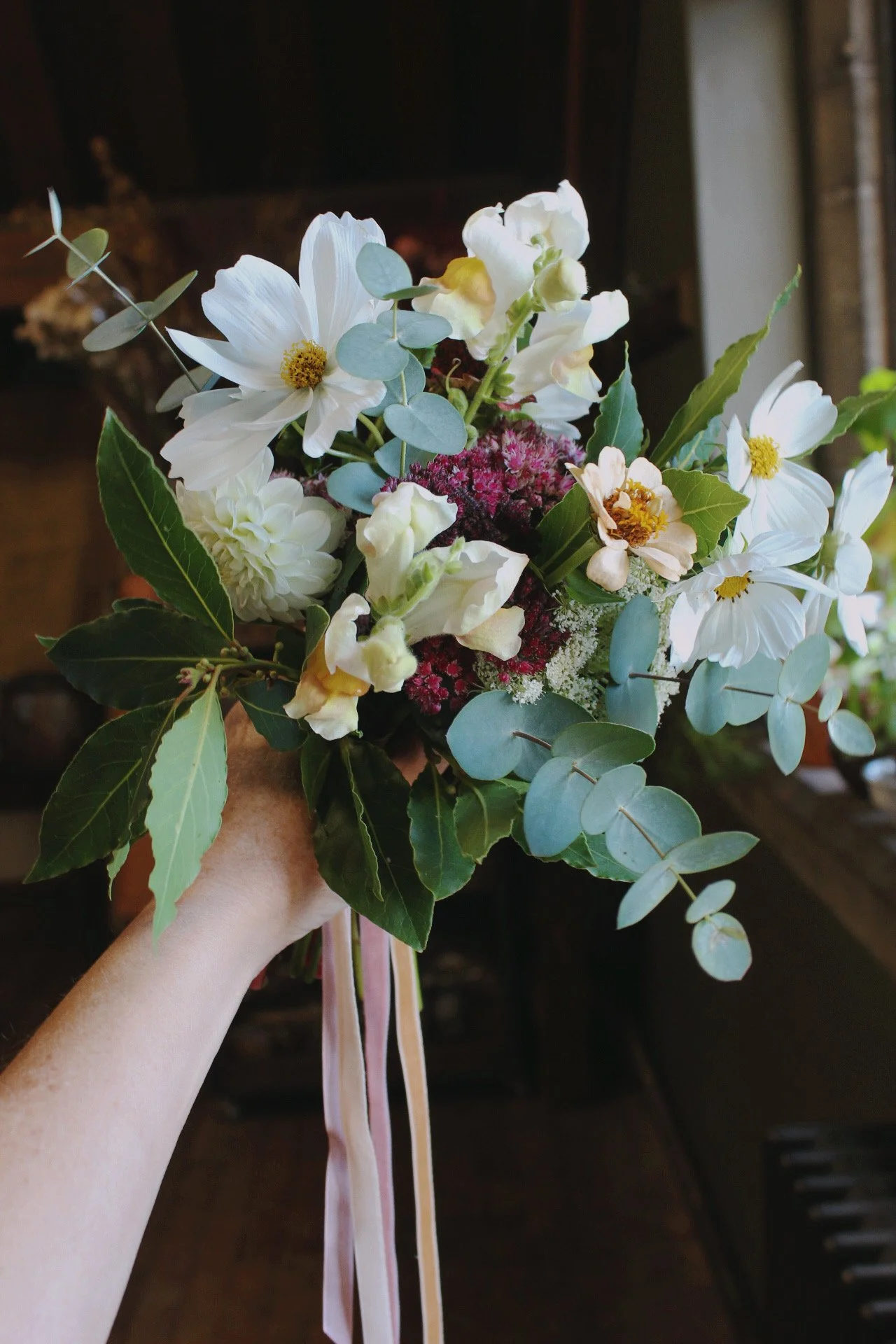 A hand holding a flower girl, wedding bouquet of white flowers, green leaves, and pink elements, tied with pink and white ribbons.