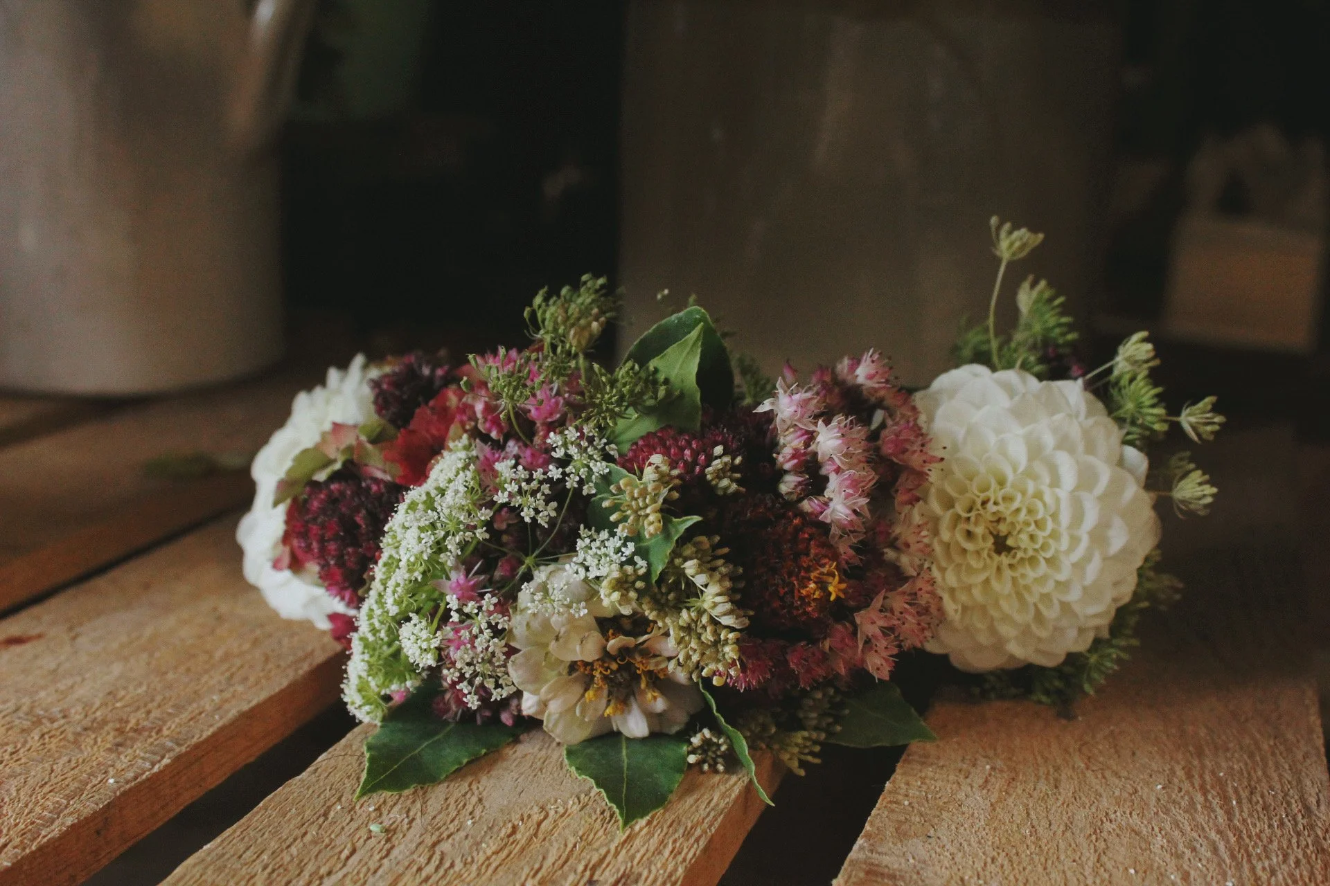 A freshly arranged dog colour of mixed flowers including white hydrangeas, pink and dark red roses, and greenery, placed on a wooden surface indoors for a wedding.