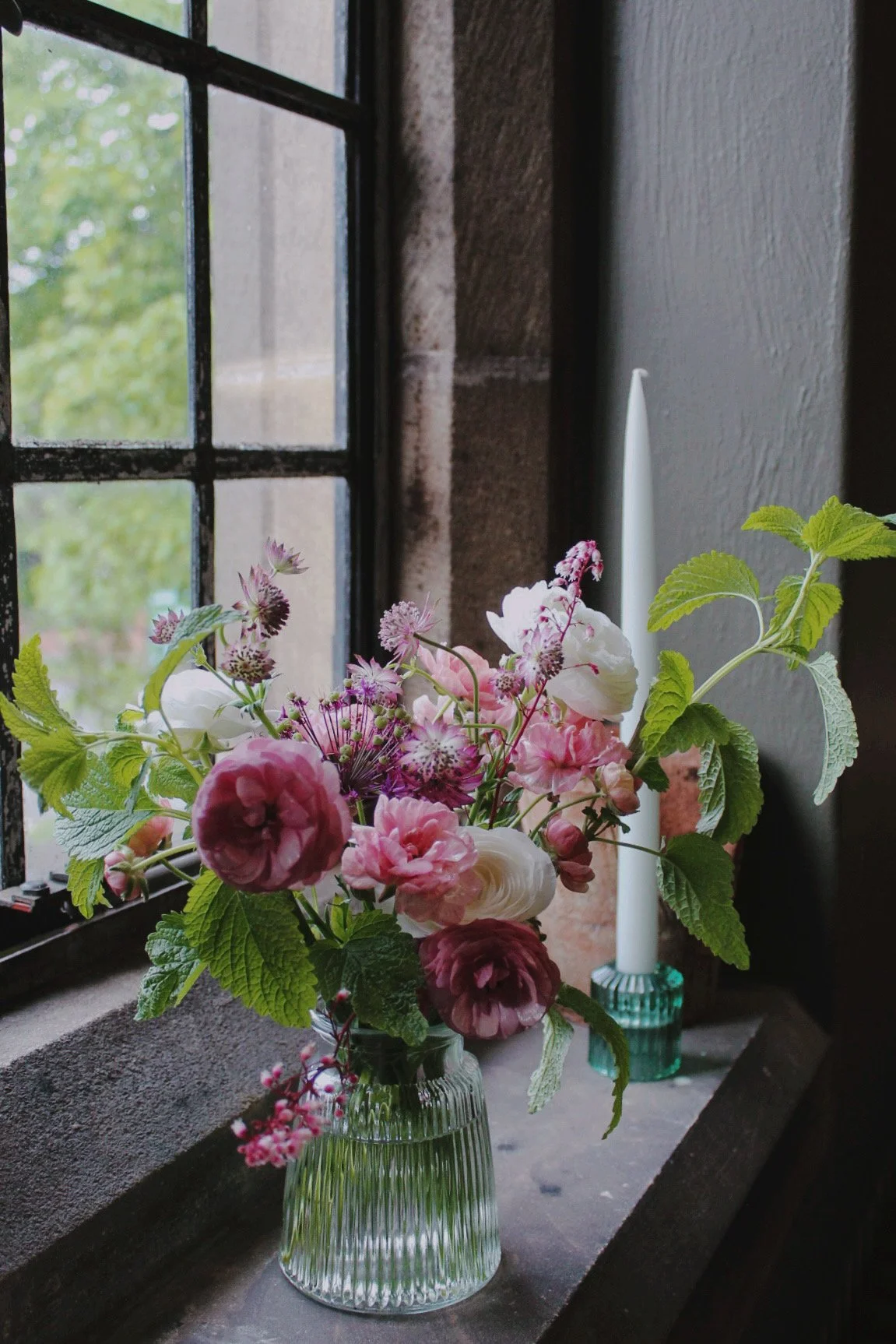Ribbed glass cocktail centrepiece filled with seasonal pink and white flowers, styled with a taper candle beside a window for a wedding setting.