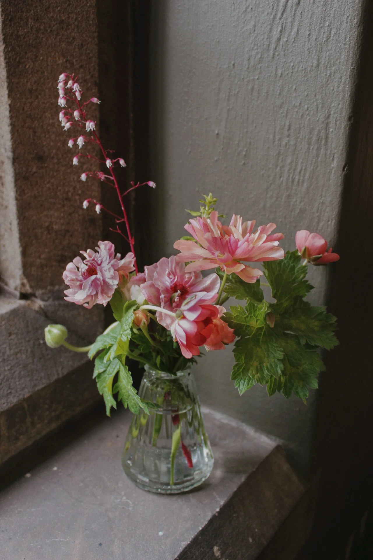 Fluted glass bud vase filled with pink and coral seasonal flowers and fresh green foliage, styled on a windowsill.