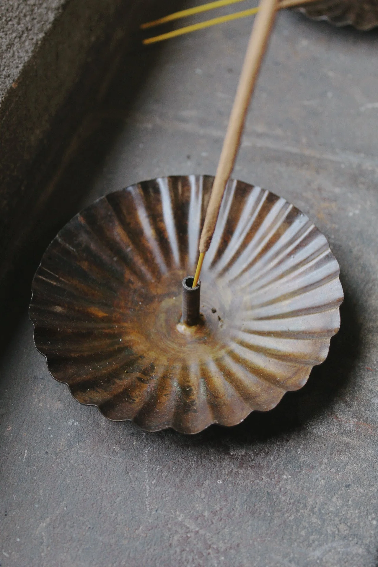Scalloped metal incense holder with an aged finish, shown with a lit incense stick, incense sticks and terracotta pots.
