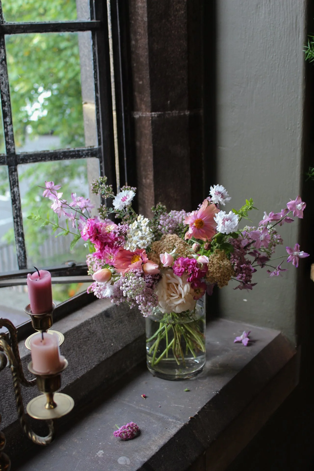 Glass cocktail centrepiece filled with seasonal wedding flowers styled beside candles on a stone windowsill.