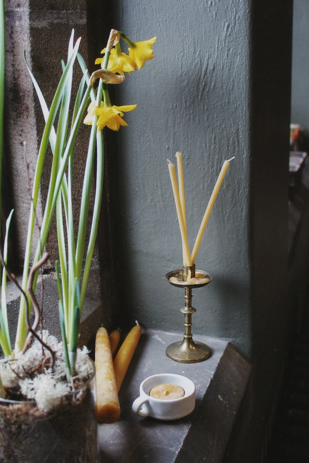 Small white ceramic candle dish with a beeswax tea light beside daffodils and candles.