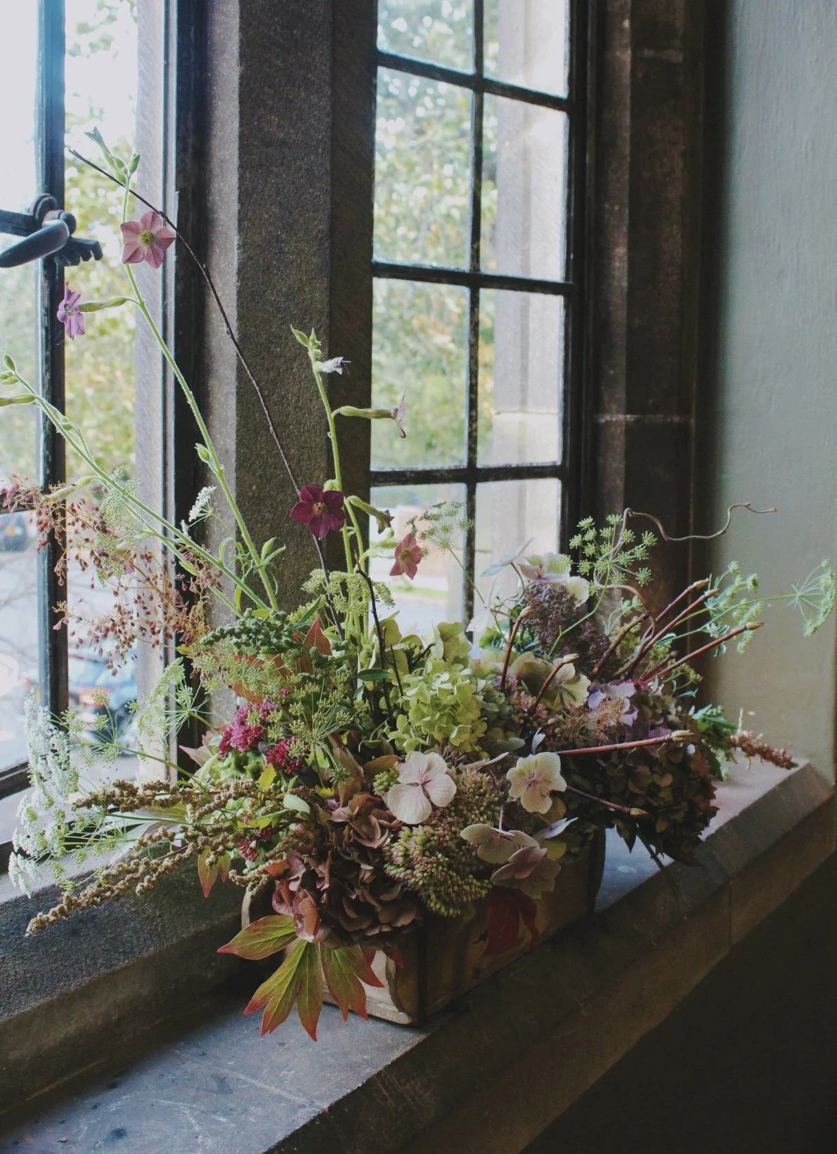 A wedding tablescape trough flower arrangement with various pink, white, and green flowers and foliage on a window sill, next to a black window frame and glass panes.
