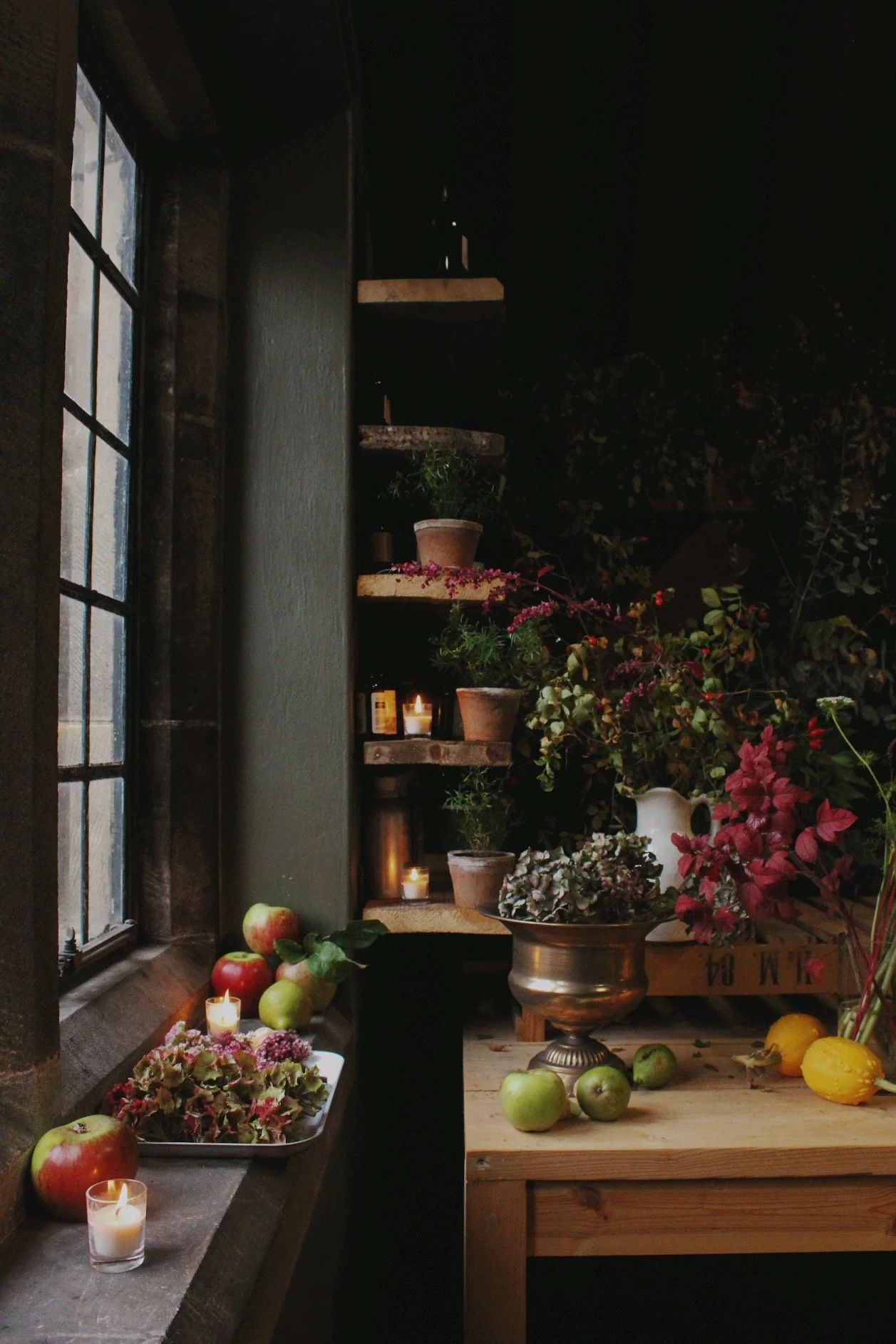 A rustic windowsill with lit candles, apples, and potted plants, vases of fresh autumnal flowers, next to a wooden table holding more apples and a large bouquet of flowers, within a cozy, dimly lit space.