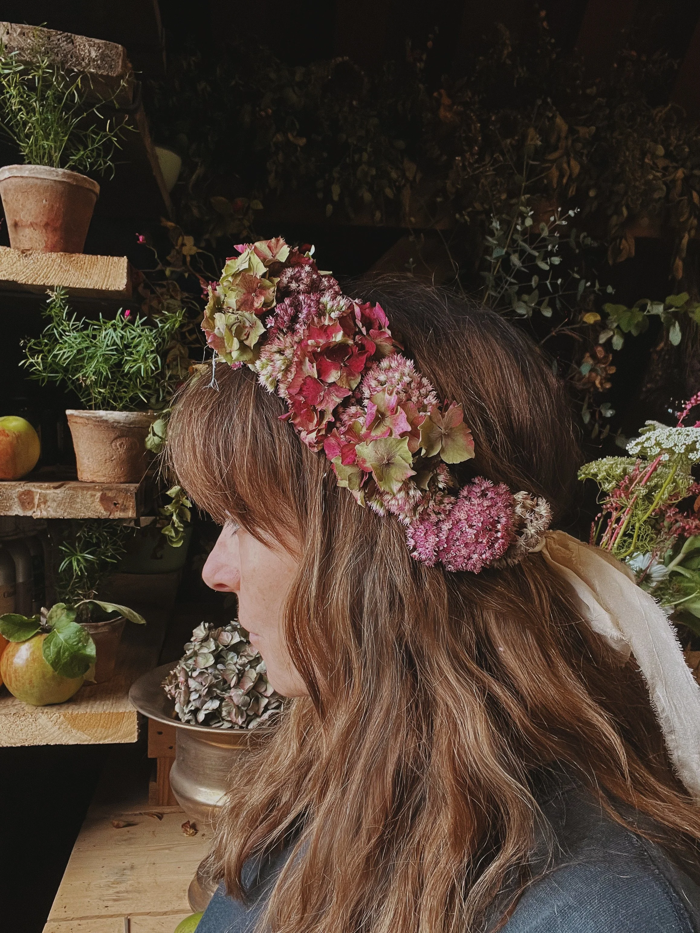 A woman with long brown hair wearing a flower crown with silk ribbon made of autumn pink and green flowers. She is surrounded by potted plants and flowers on shelves in a studio.