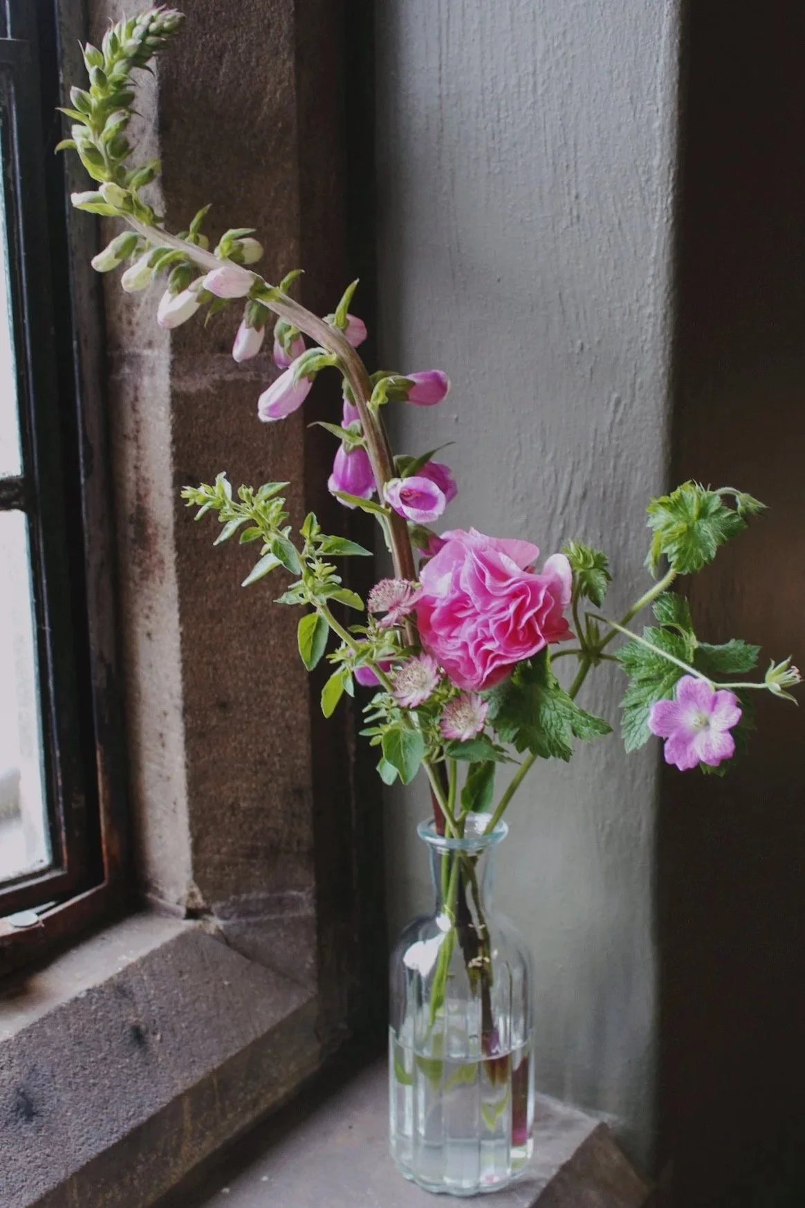 Ribbed glass bottle bud vase filled with pink seasonal flowers, foxglove and delicate foliage, styled on a stone windowsill.