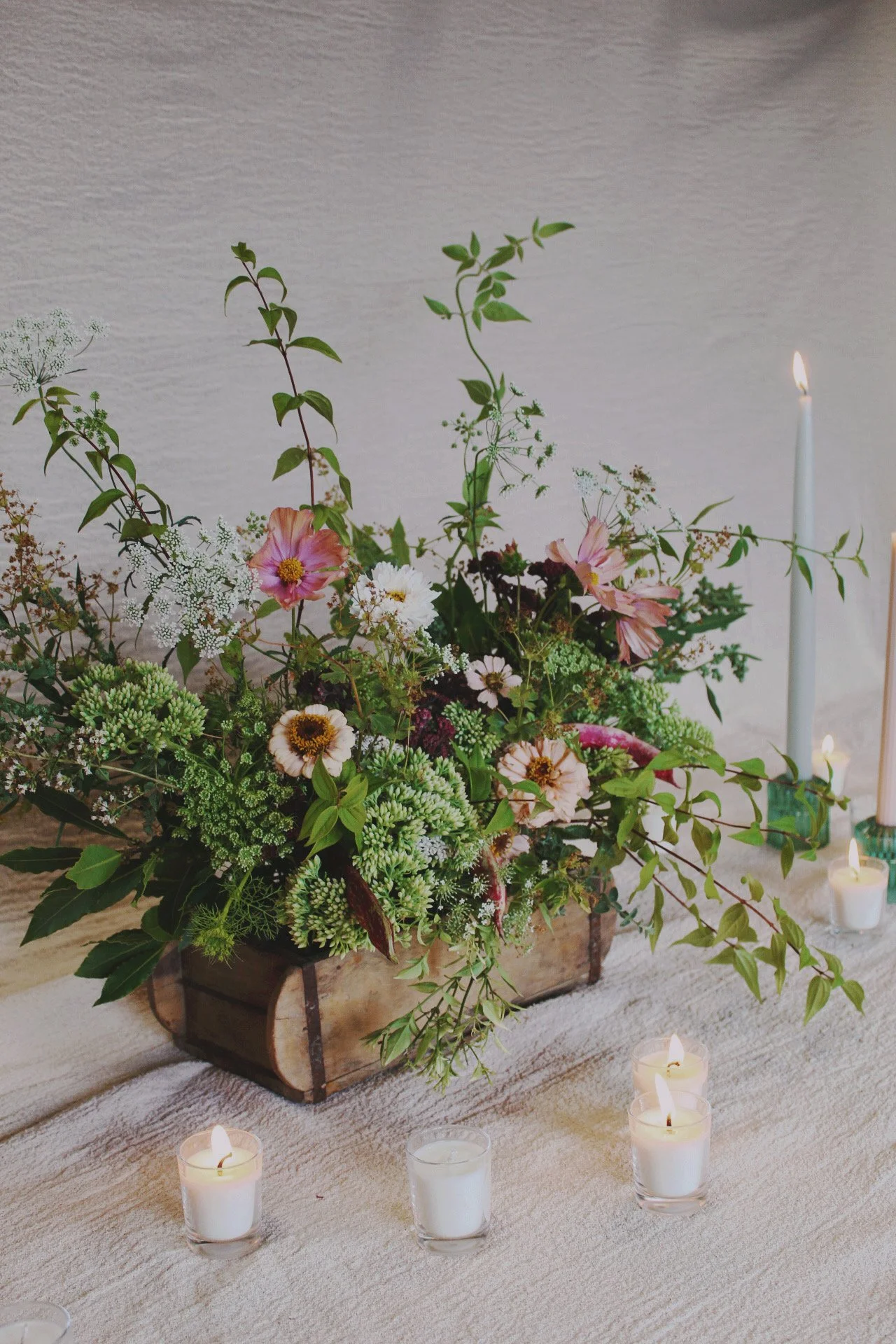 A wedding tablescape trough floral arrangement with pink, white, and green flowers and greenery in a wooden container, surrounded by lit candles on a white textured tablecloth.