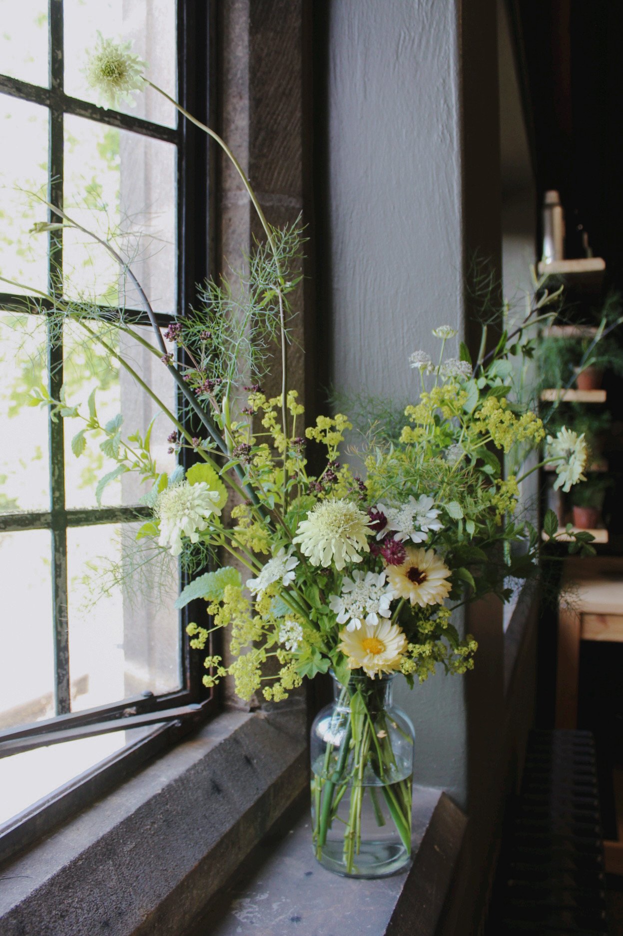 A wedding arrangement of assorted flowers in a glass vase sitting on a windowsill next to a window with black windowpane bars, with a gray wall in the background.