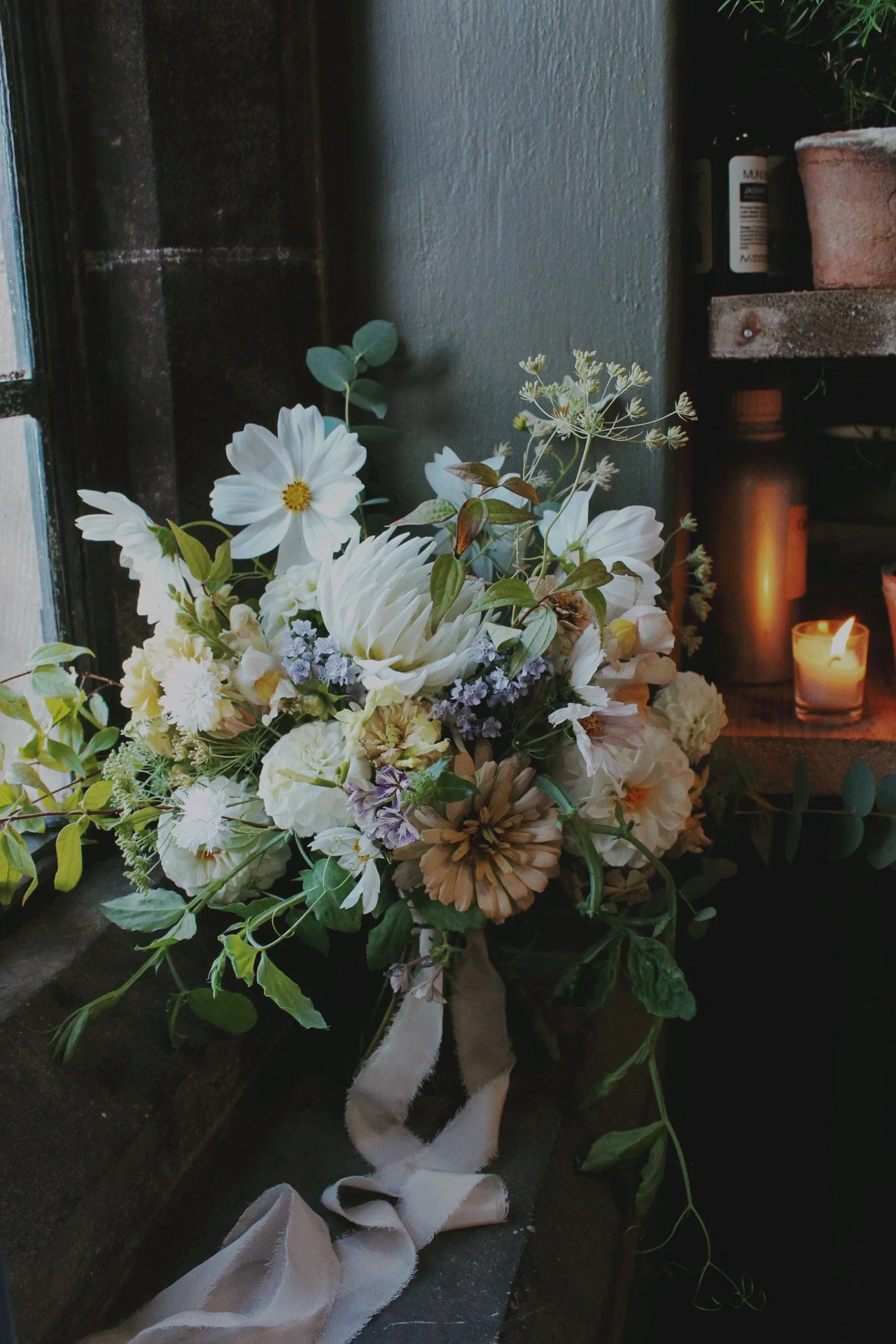 A bridal bouquet of white and soft neutral seasonal flowers with delicate greenery, displayed beside a candle in the studio.