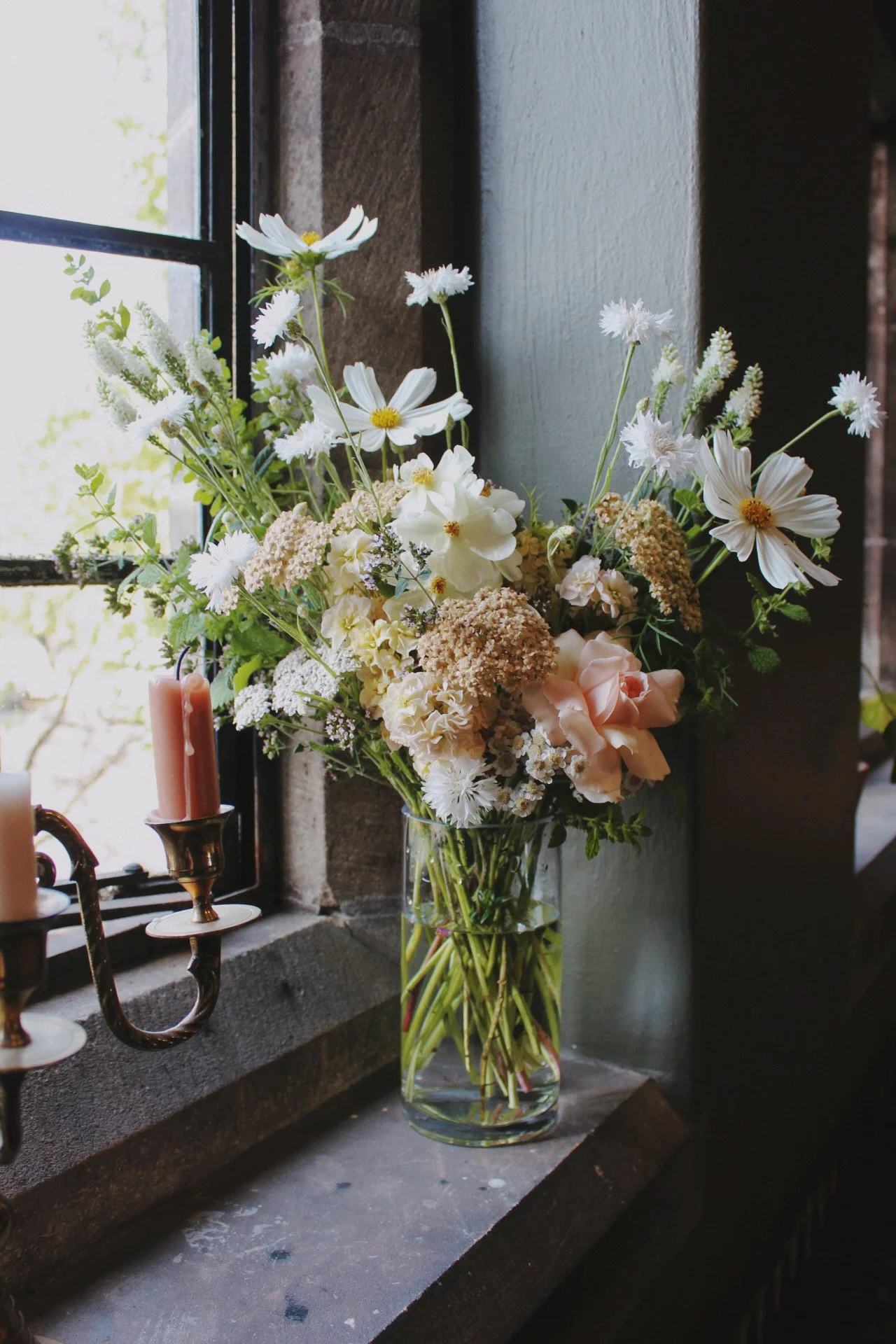 Classic glass centrepiece filled with seasonal white and blush flowers, foliage and delicate meadow flowers beside a stone window.
