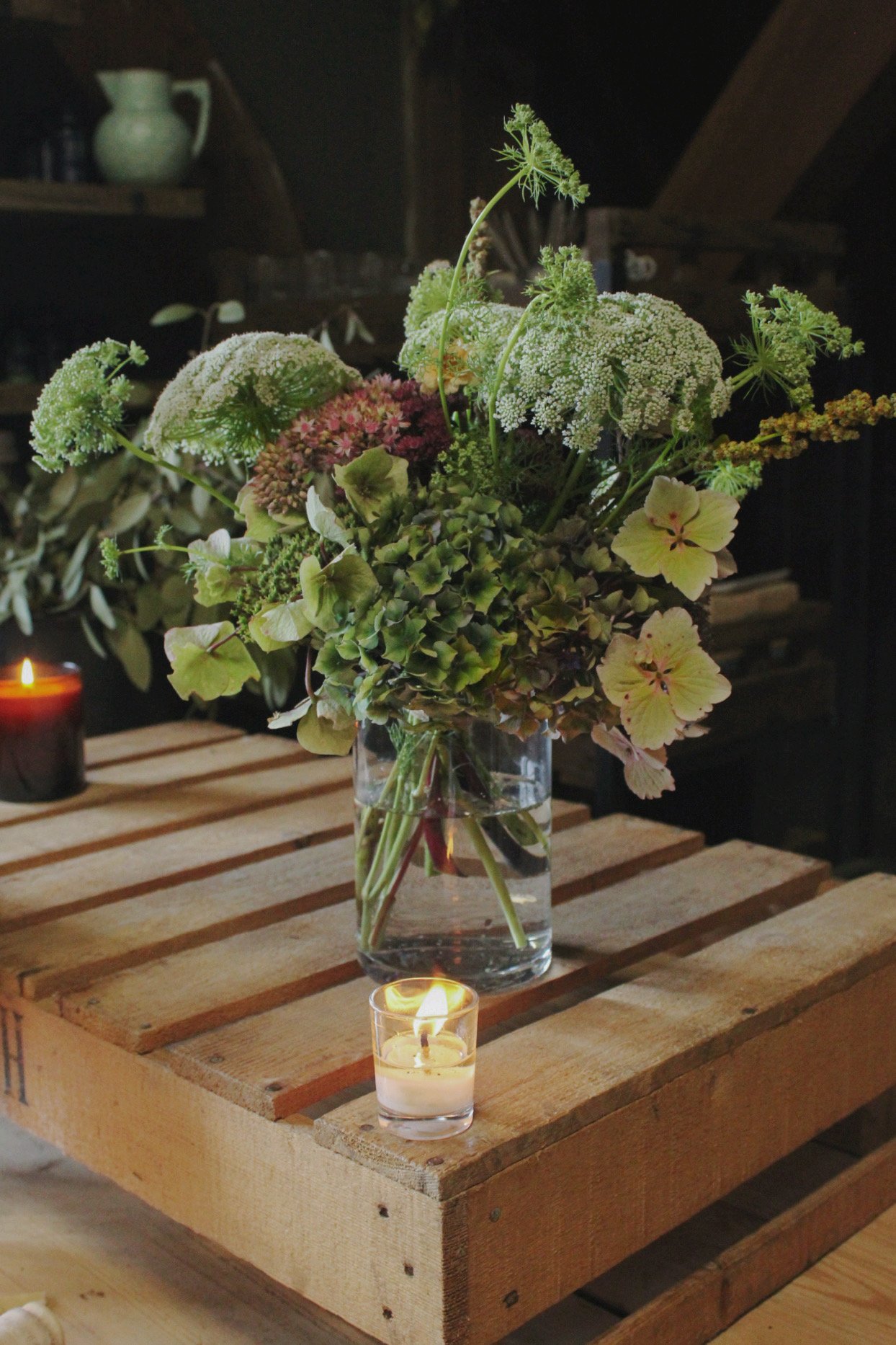 A bouquet of mixed flowers in a glass vase with water on a wooden table, surrounded by lit candles in small glass holders, in a cozy indoor setting.
