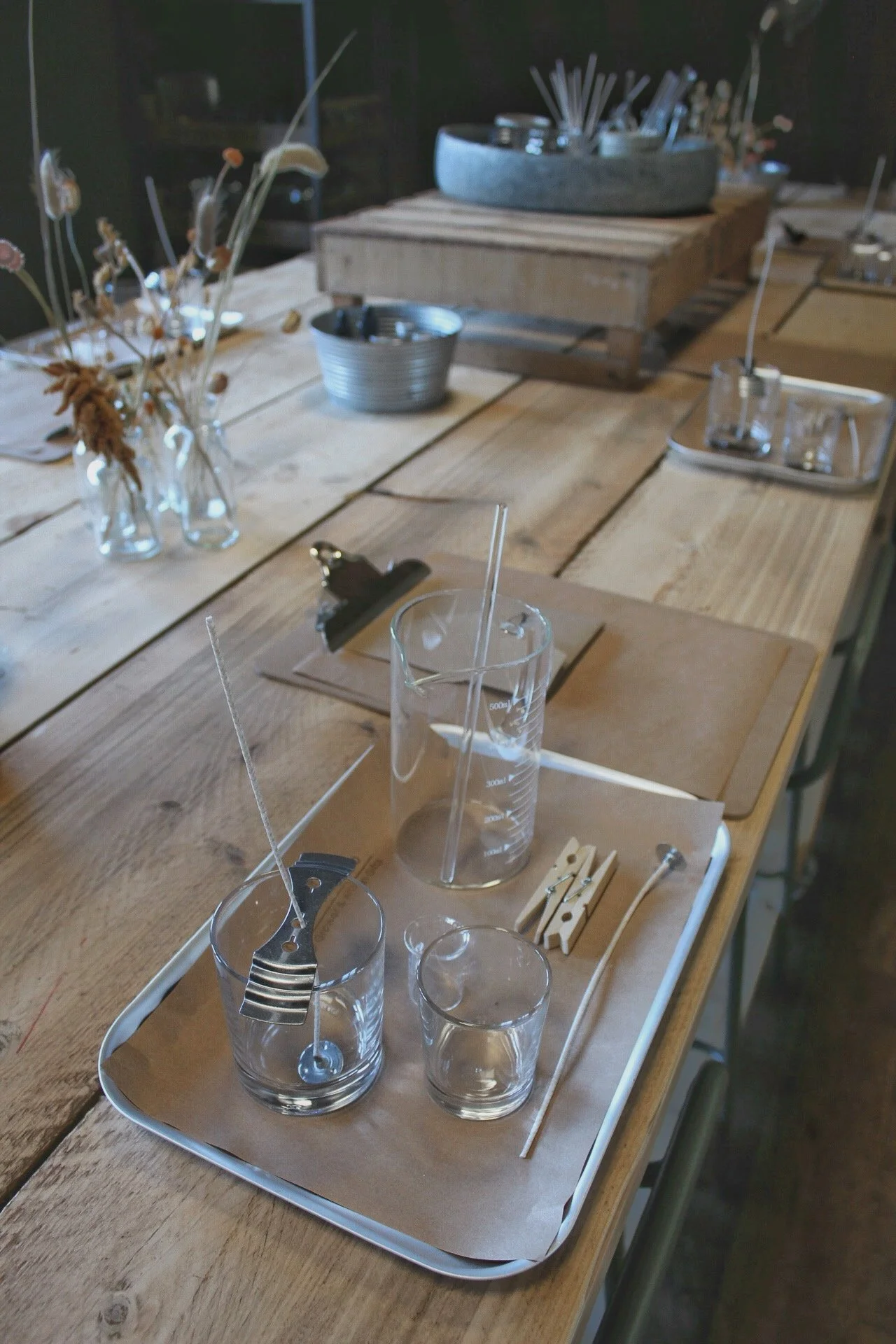 A wooden table set with glass beakers, straws, and clothespins, with dried flowers in small glass bottles in the background.