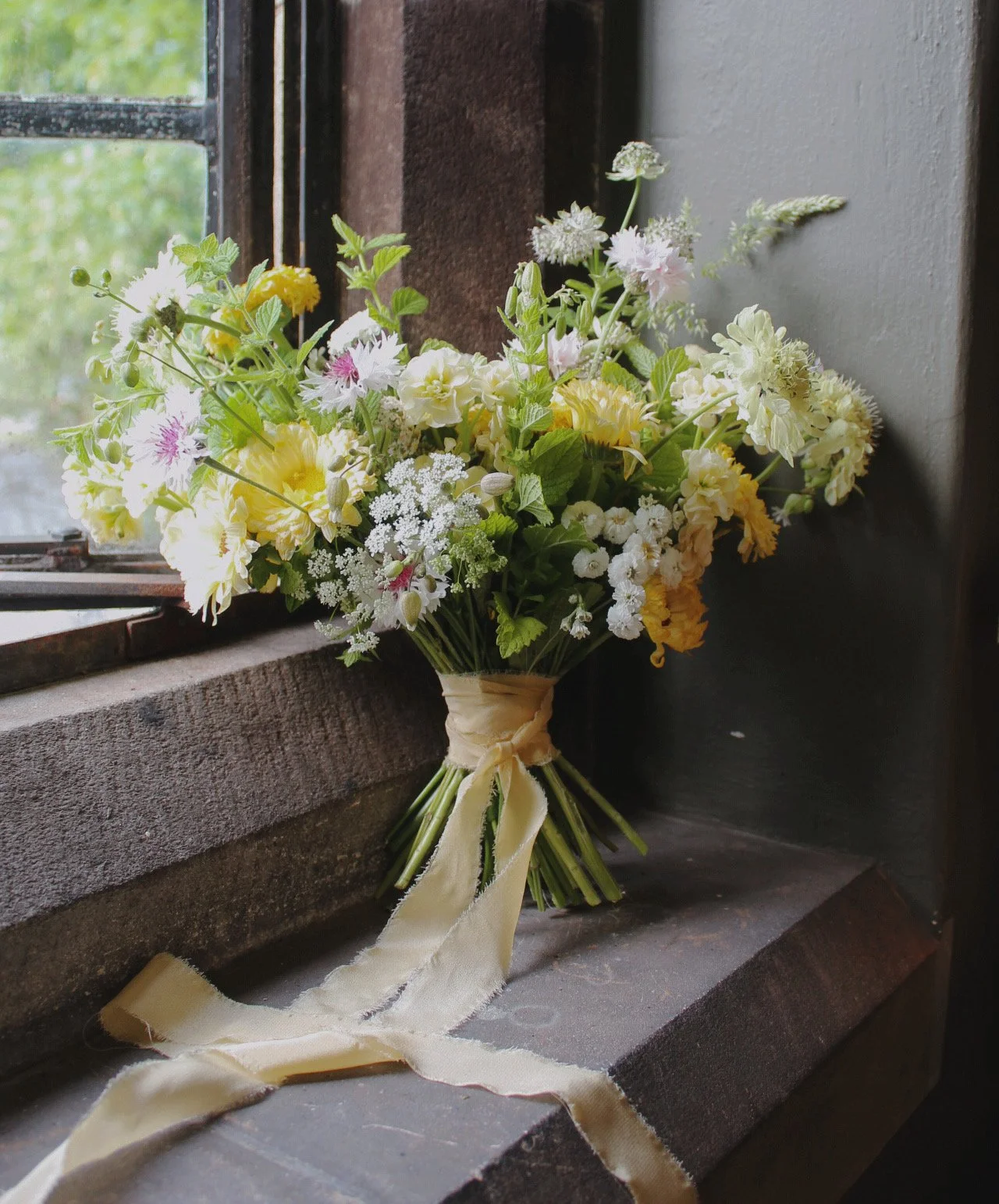 Edit bridal bouquet with soft yellow, white and pale pink flowers, fresh foliage and flowing silk ribbon.