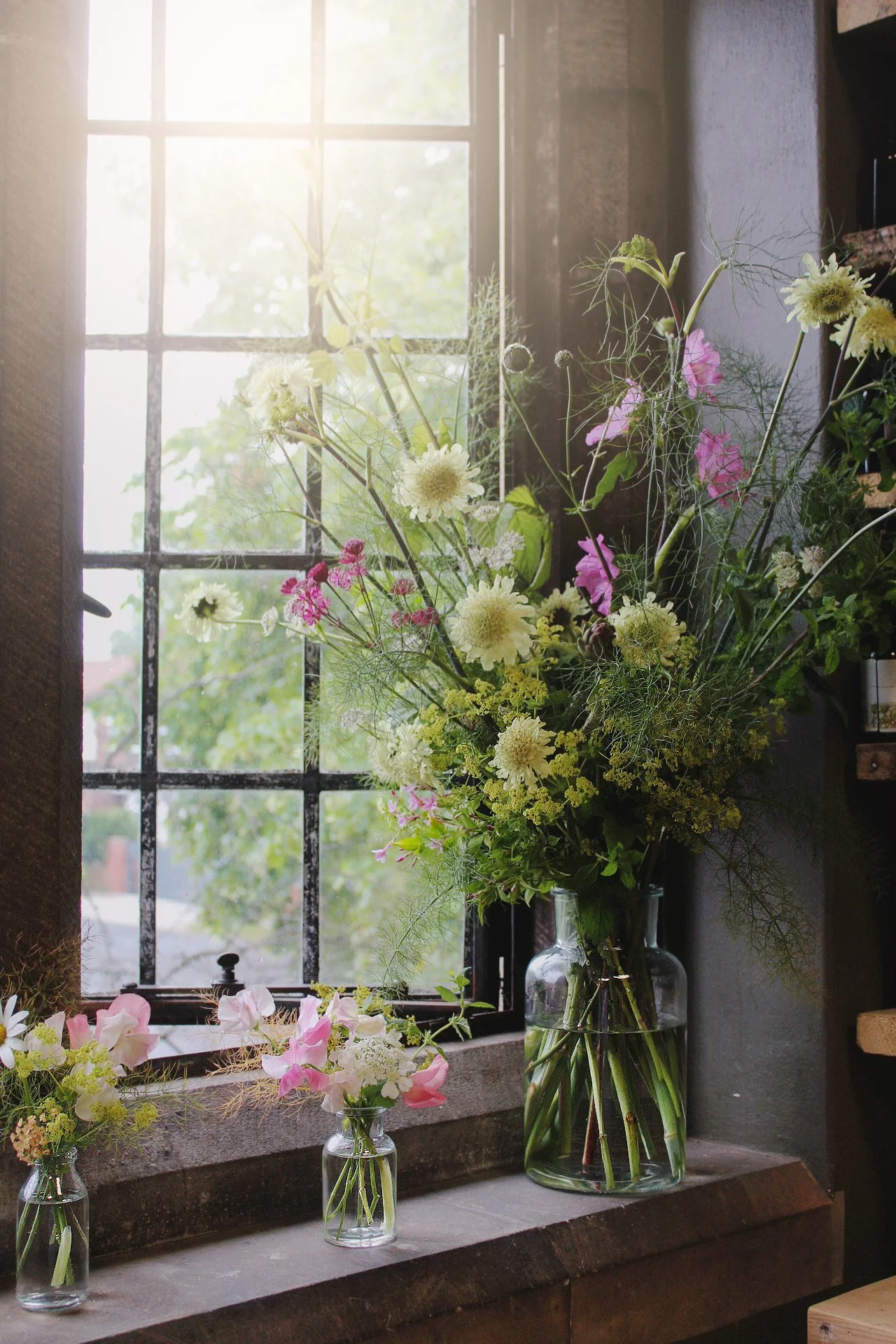 Tall glass statement vase filled with an abundant, airy arrangement of pink, white and green seasonal flowers beside a window.