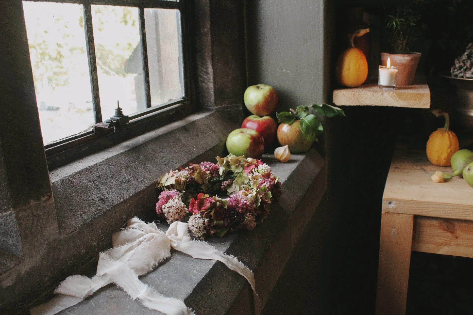 A windowsill holding three apples, and autumnal fruit with a wedding flower crown. To the right, a wooden shelf with a pumpkin, a lit candle, and potted plants.