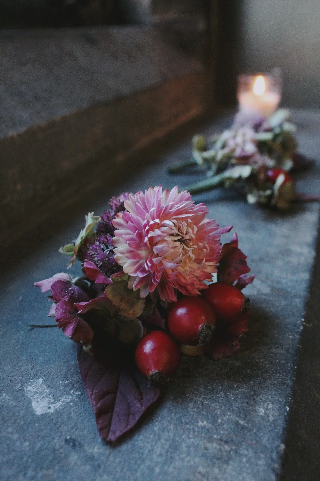 A floral arrangement with pink and purple flowers, red berries, and purple leaves on a gray surface, with a candle and a wooden surface in the background for a grooms buttonhole.