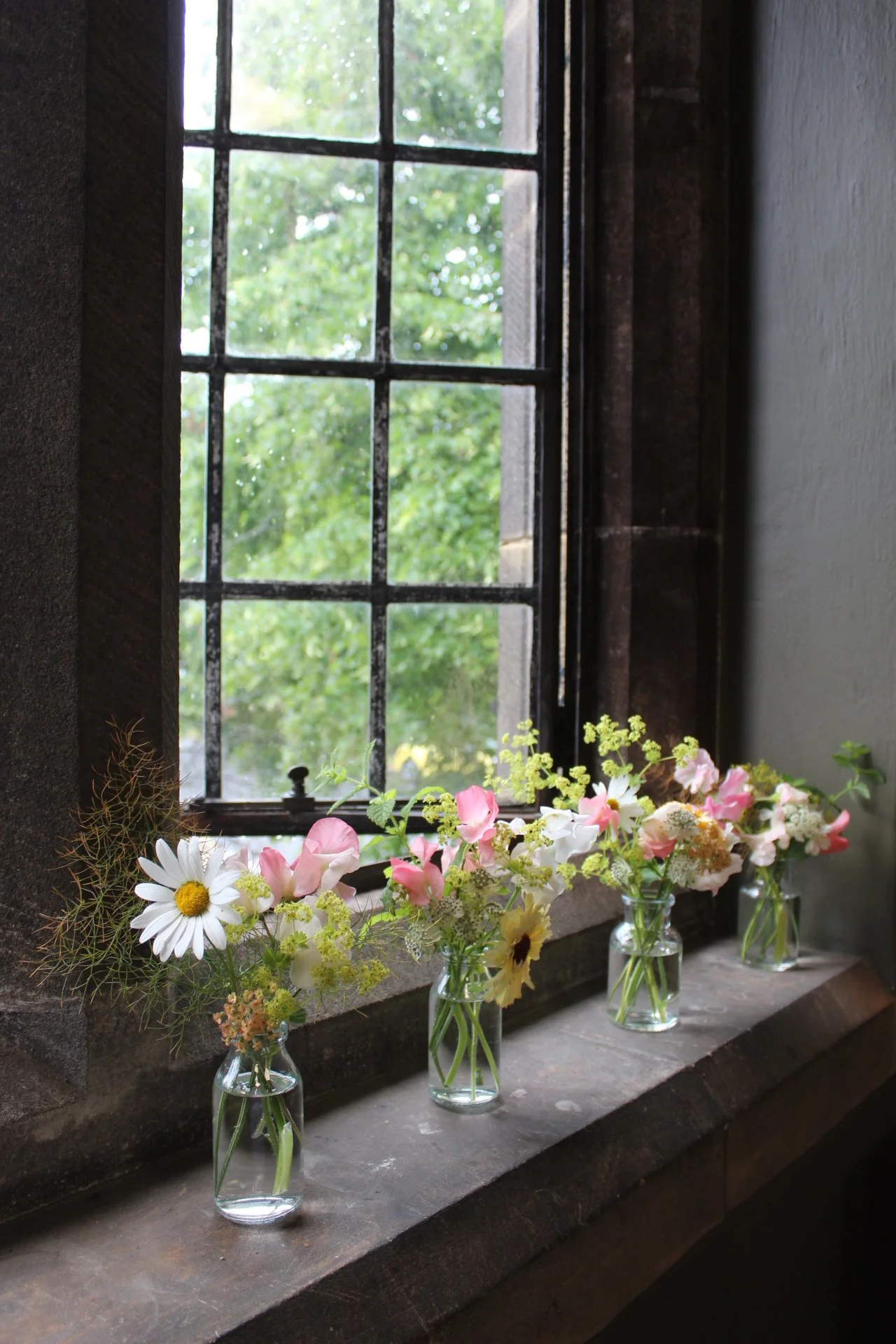 Three bud vases containing wedding flowers on a windowsill next to a window showing green trees outside.