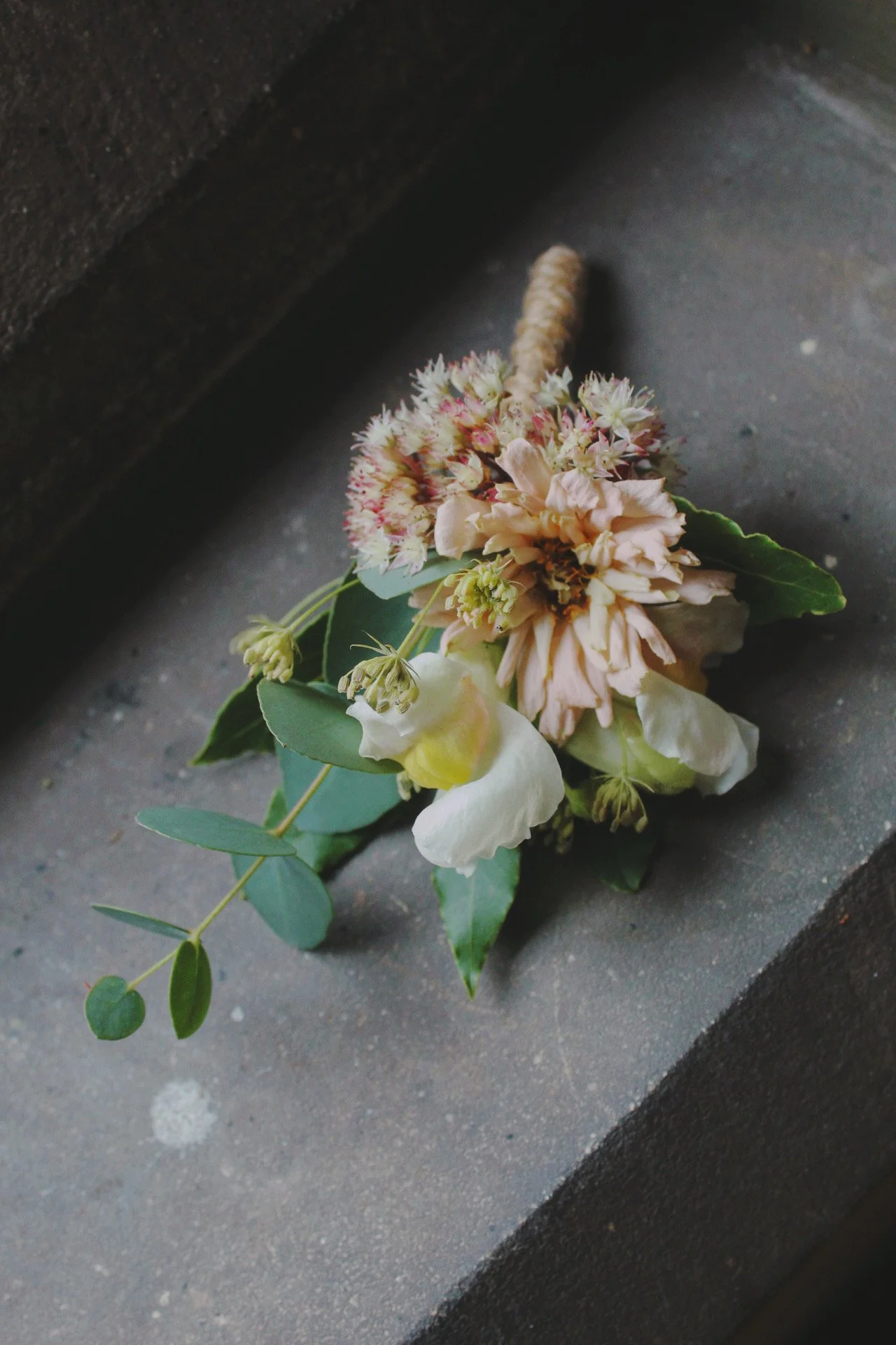 A wedding party buttonhole of mixed flowers with green leaves, lying on a dark, textured surface.