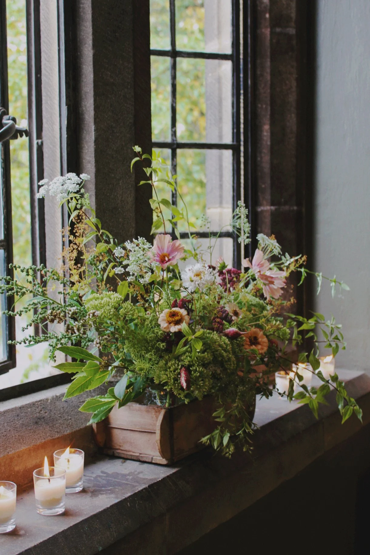 Wedding tablescape trough filled with seasonal flowers, foliage and trailing greenery, styled with candles beside a window.