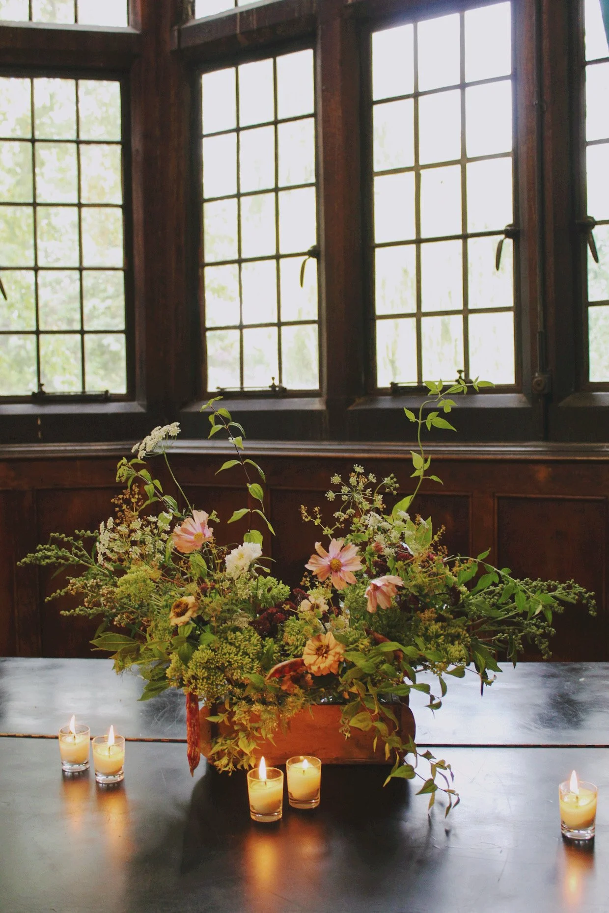 Wedding tablescape trough filled with seasonal flowers, foliage and trailing greenery, styled with candles on a long dining table.