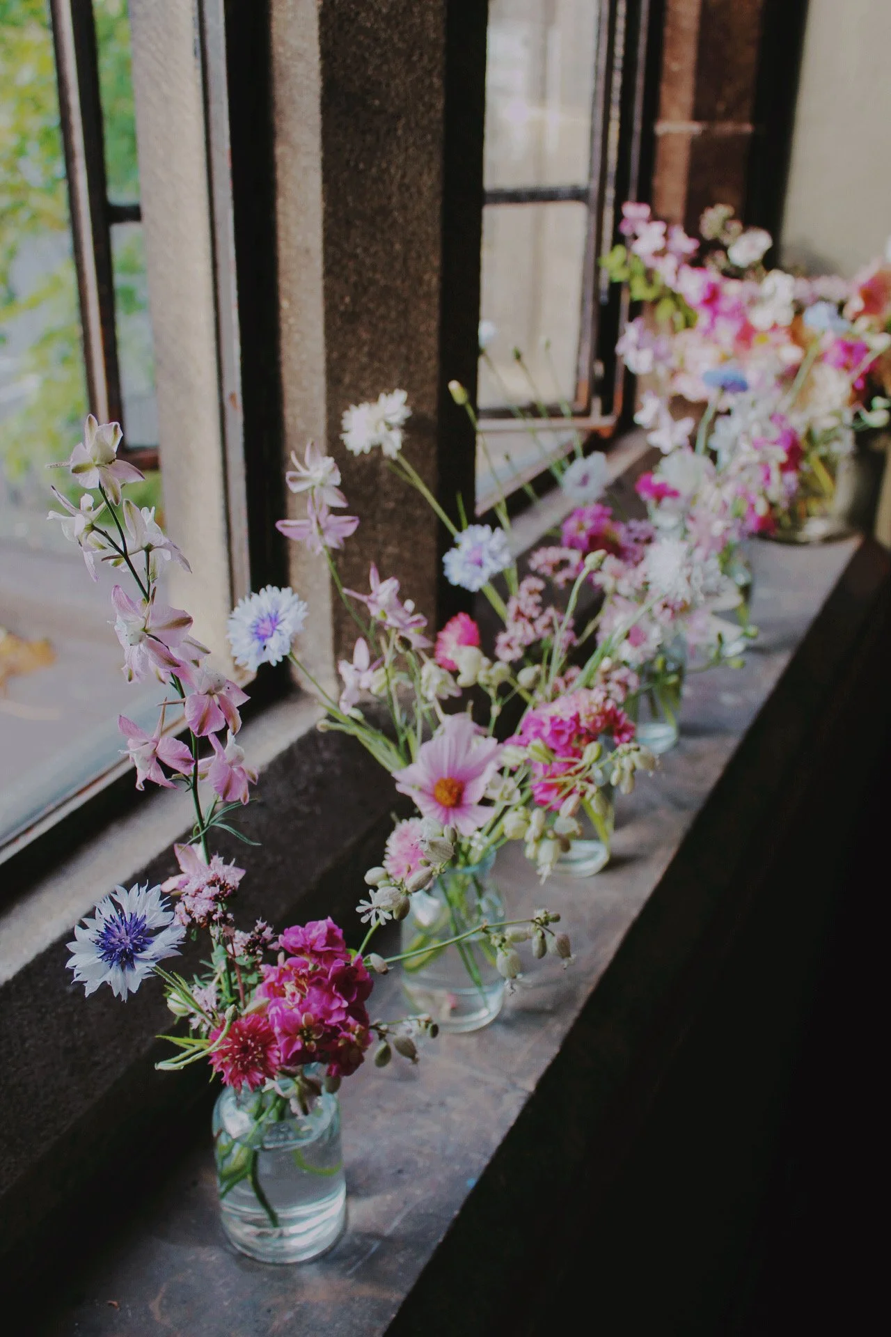 Signature bud vases styled along a windowsill with delicate seasonal flowers in soft pink, white and blue tones.