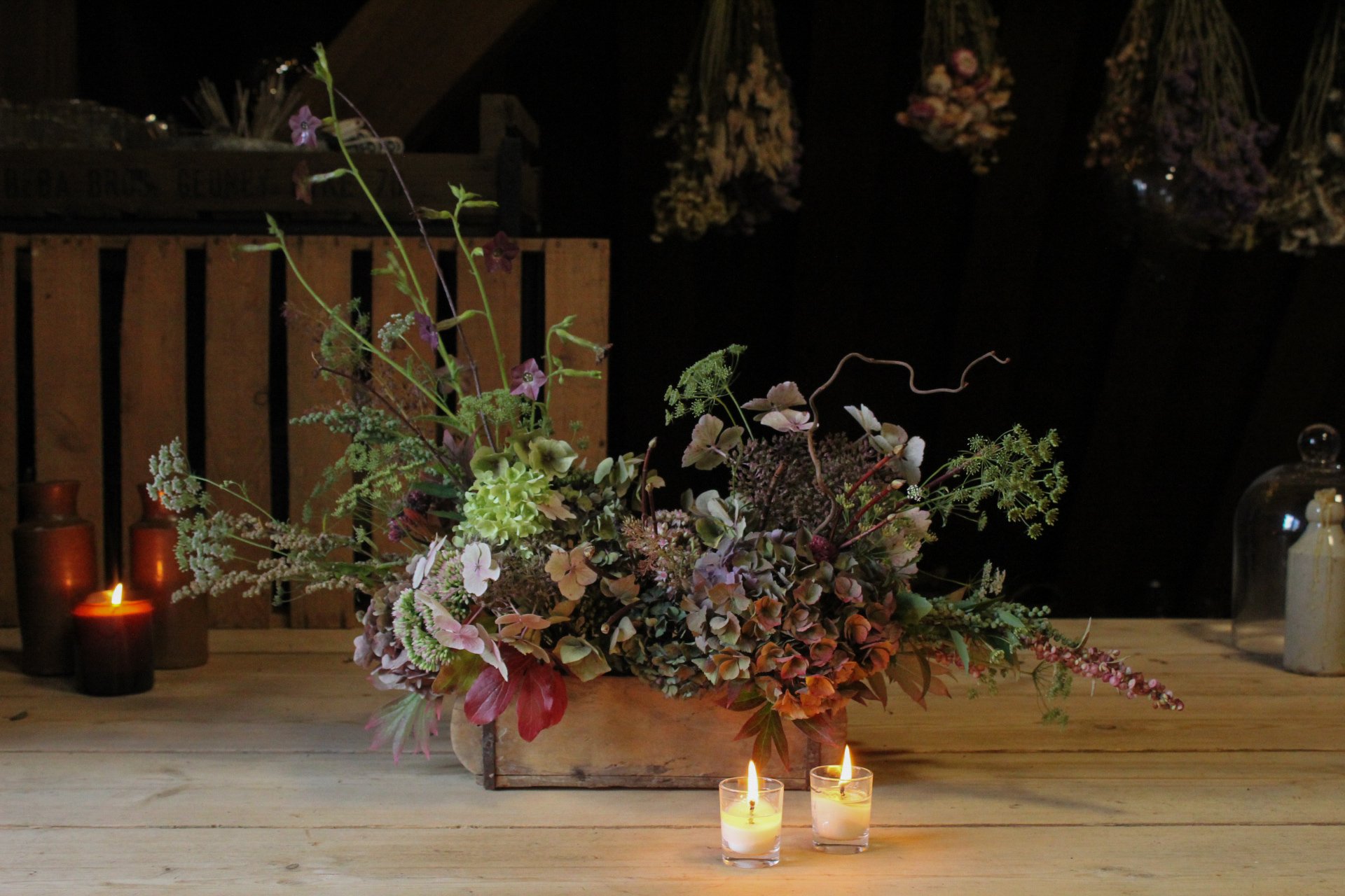A garden led floral arrangement in a wooden planter surrounded by glowing candles and hanging dried flowers