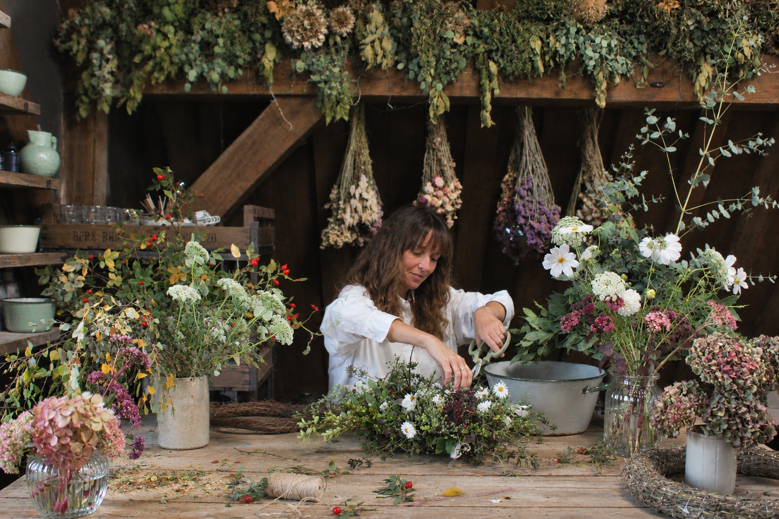 The florist behind The Wild Potager Co. arranging seasonal flowers at her studio worktable, surrounded by gathered blooms and dried flowers.