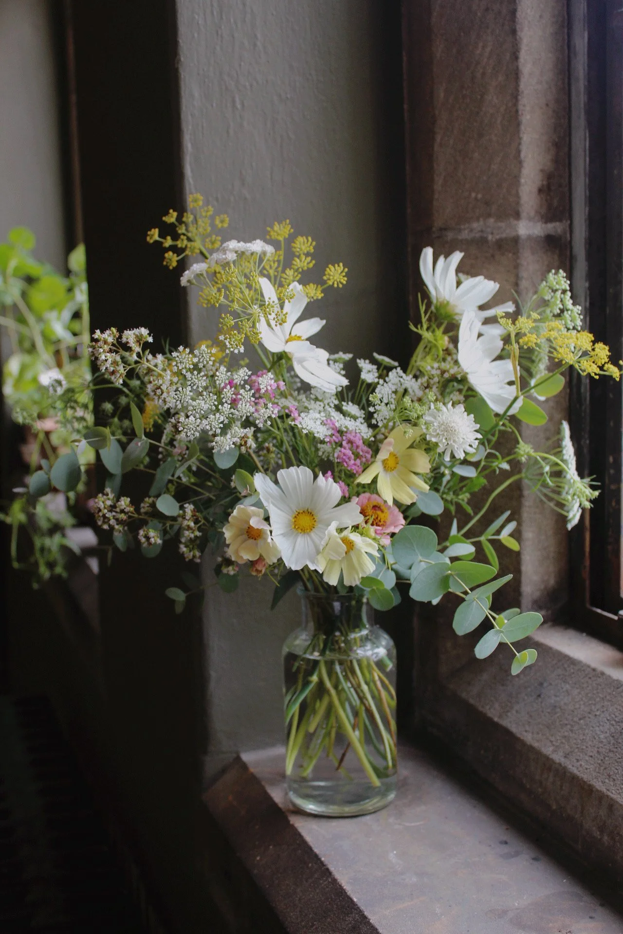 Compact glass vase filled with a loose, natural arrangement of white and soft pastel seasonal flowers on a stone windowsill.