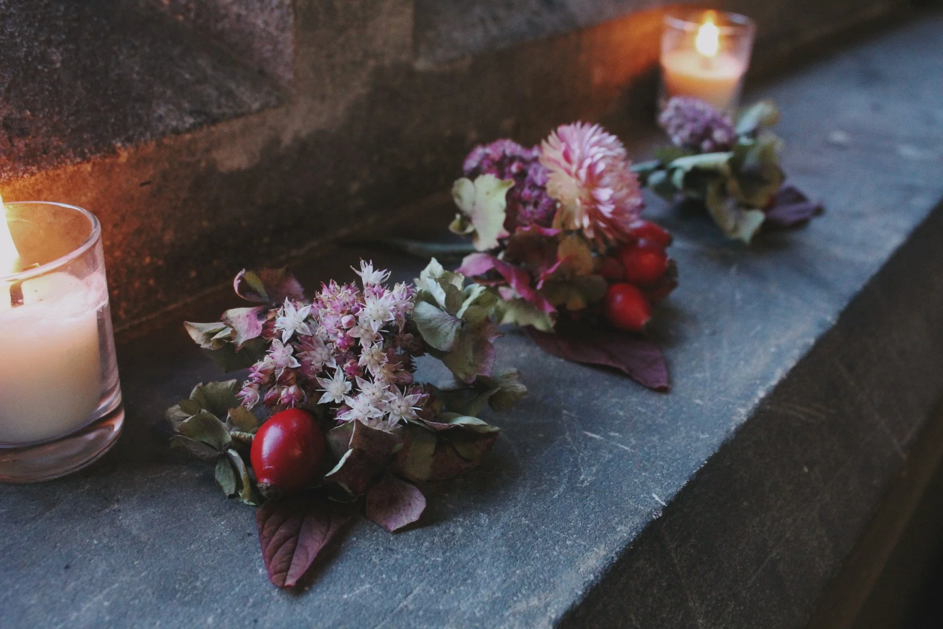 Votive candles with wedding party buttonhole flowers and red berries on a dark stone surface beside a brick wall.