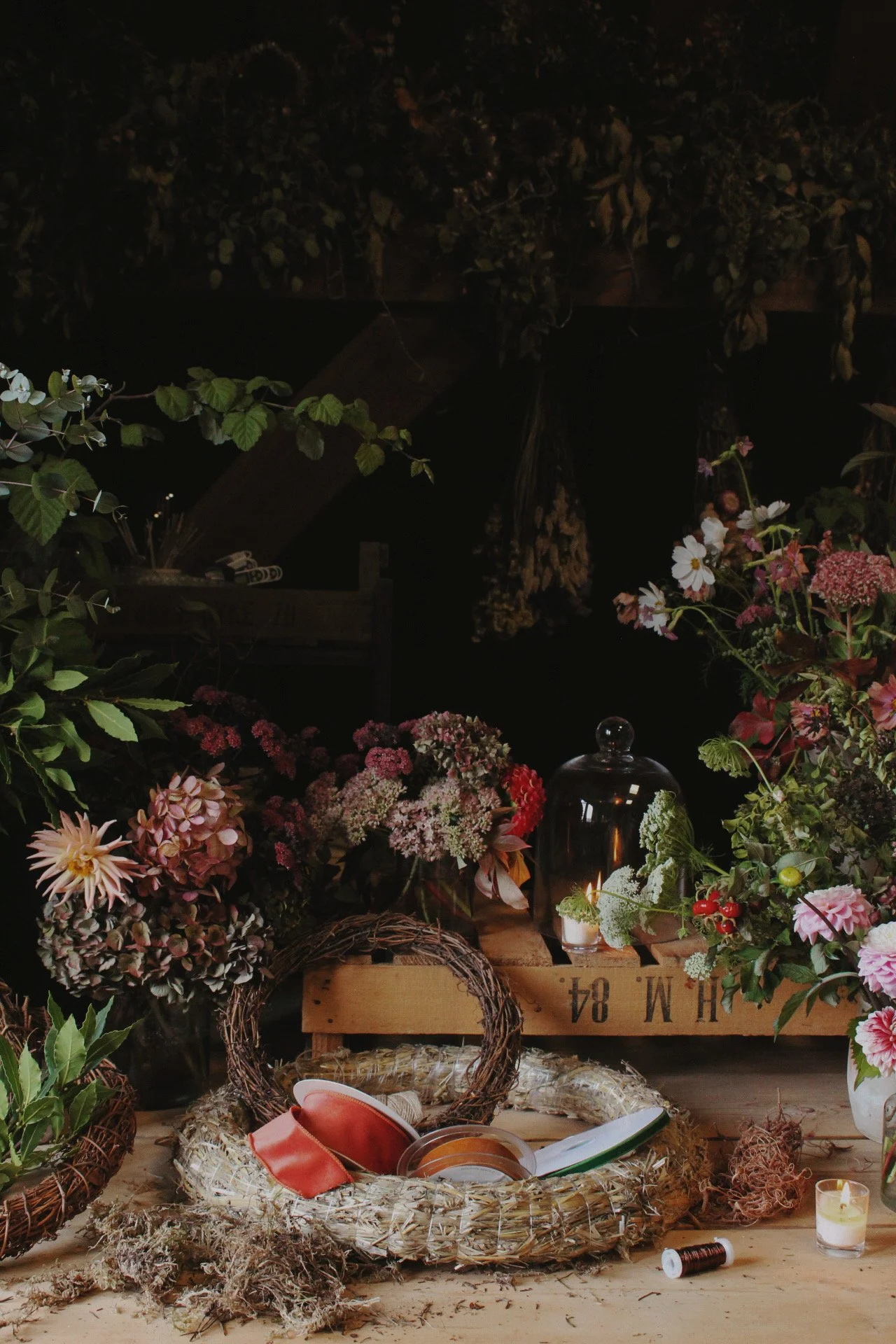 A display of various flowers, plants, candles, and wreath materials arranged on a table for a floral arrangement workshop. There are flowers in vases, a candle, a spool of thread, and decorative wreaths.