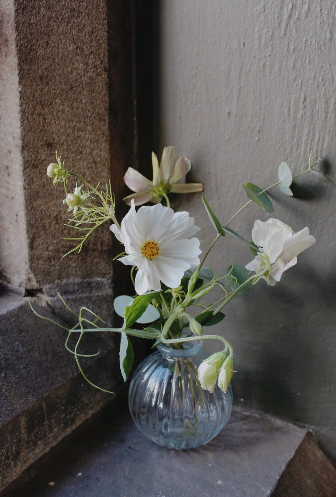 Round glass bud vase filled with delicate white seasonal flowers, eucalyptus and trailing sweet pea foliage, styled on a windowsill.