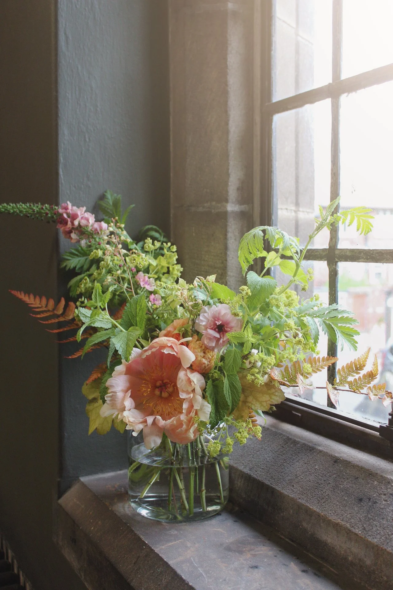 Compact glass vase filled with a loose, natural arrangement of white and soft pastel seasonal flowers on a stone windowsill.