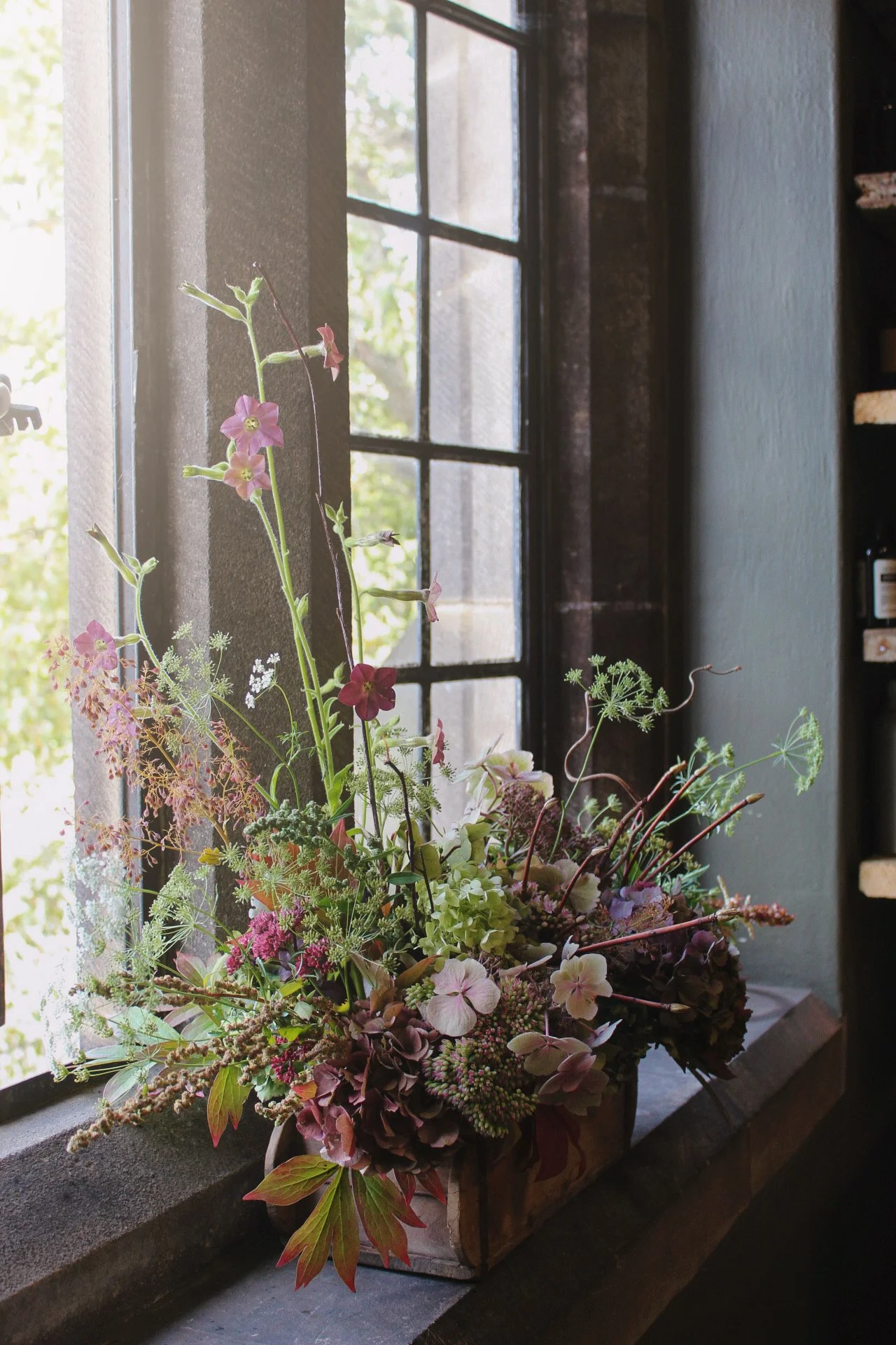 A wedding tablescape trough bouquet of pink, white, and green flowers placed on a windowsill next to a window with black grid muntins, with sunlight coming through the window.