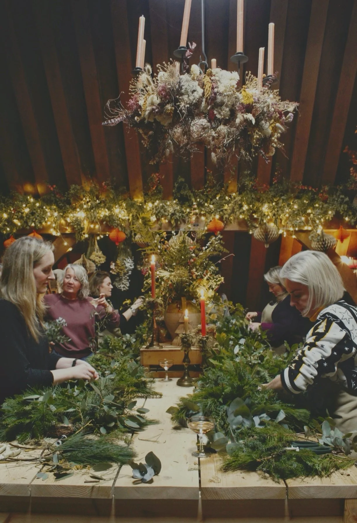 People decorating a large table with greenery and candles, with Christmas ornaments and lights hanging from the ceiling and a floral chandelier above.