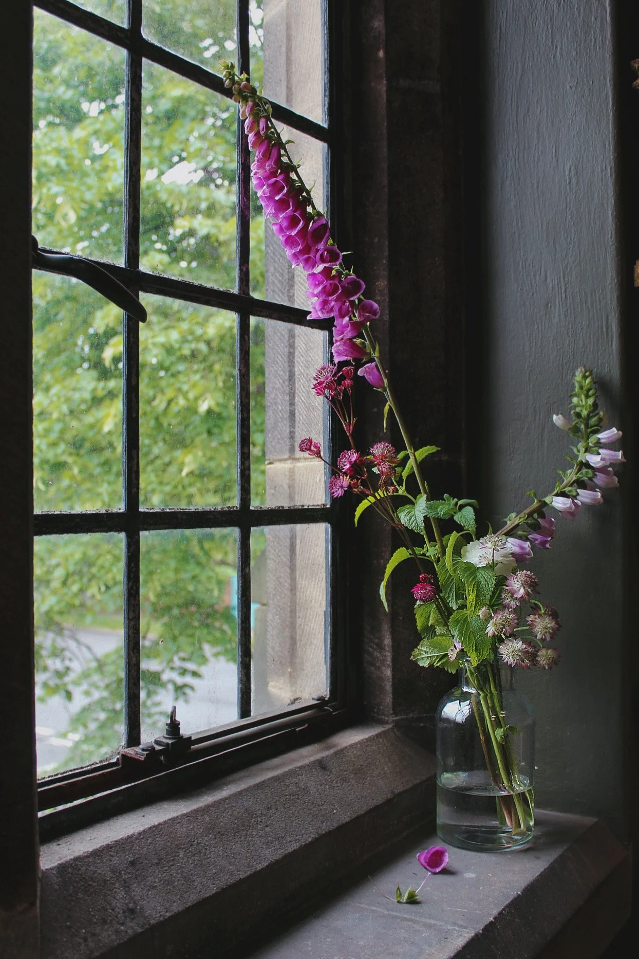 Tall glass bottle bud vase styled with elegant pink and purple seasonal flowers beside a window.