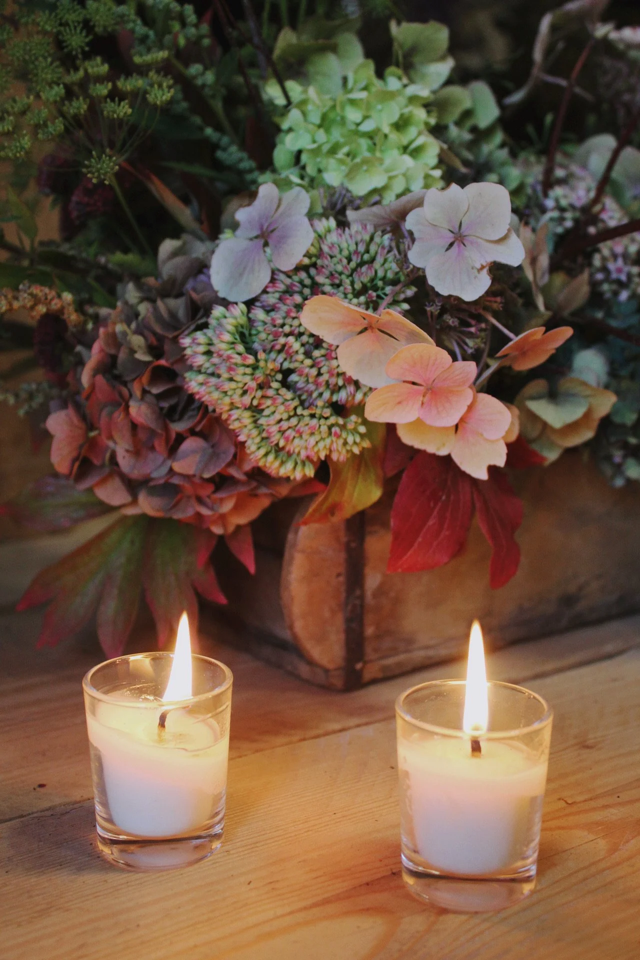 A bouquet of pastel-colored flowers, including pink, cream, and green, in a wooden container, with two lit white candles in glass holders on a wooden table in front.
