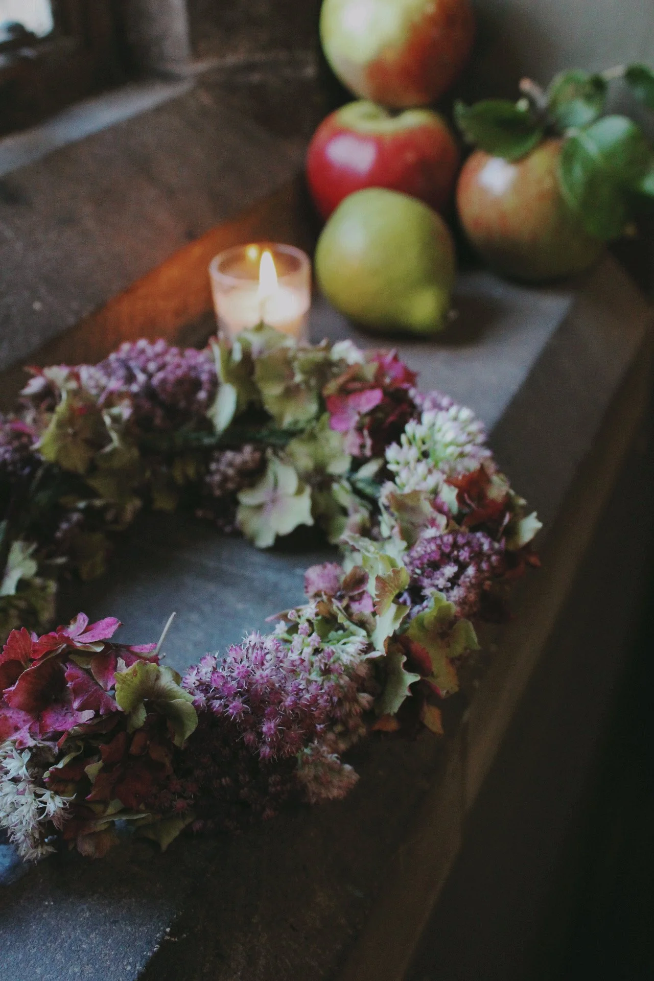 A lit candle surrounded by apples and a flower crown on a dark wooden surface.
