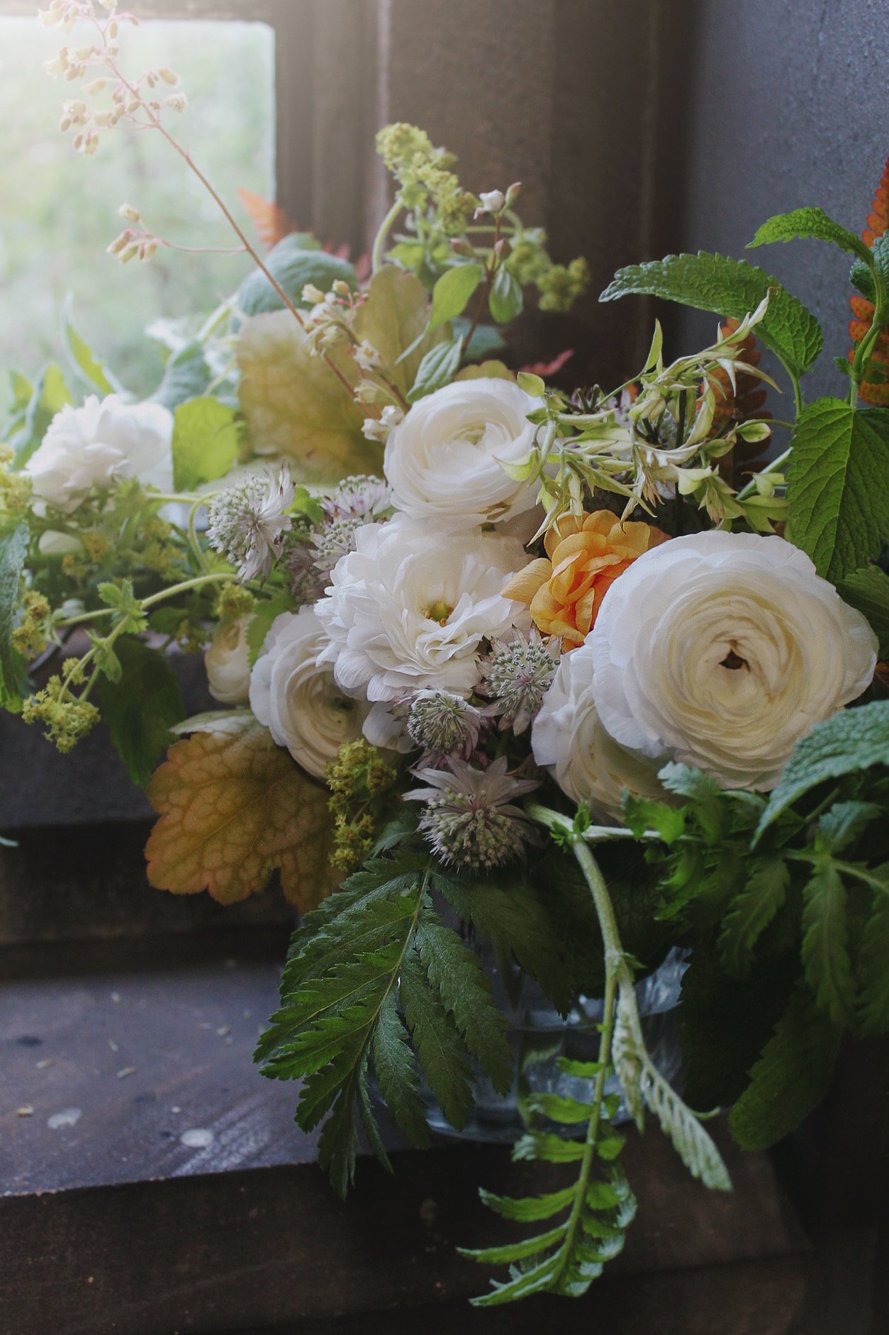 An arrangement in a vase of white ranunculus, yellow-orange flowers, assorted green leaves, and small filler flowers on a dark surface near a window with natural light.
