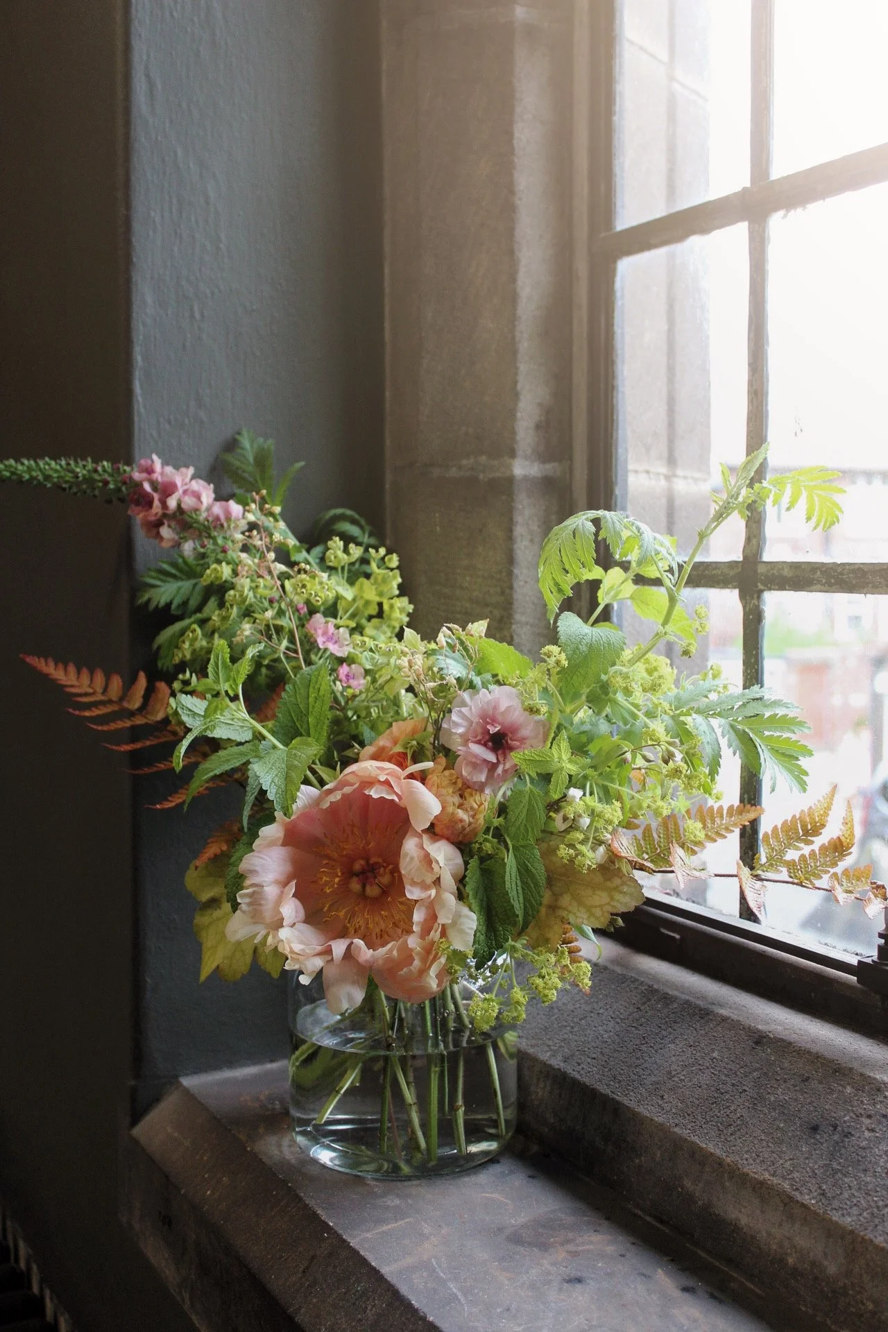 Seasonal wedding flower arrangement in a glass vase on a studio windowsill, featuring soft peach flowers, fresh green foliage and delicate flowering stems.