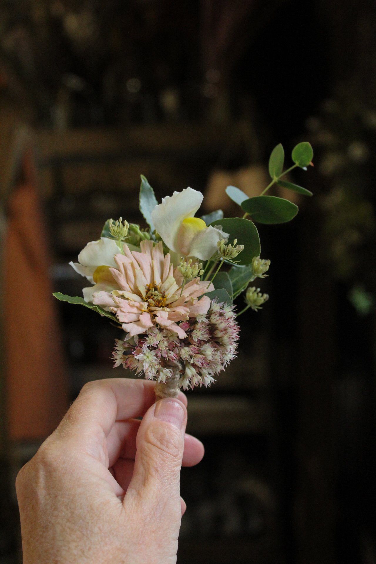 A hand holding a wedding party buttonhole, including white, pale pink, and yellow blossoms with green leaves, against a dark background.
