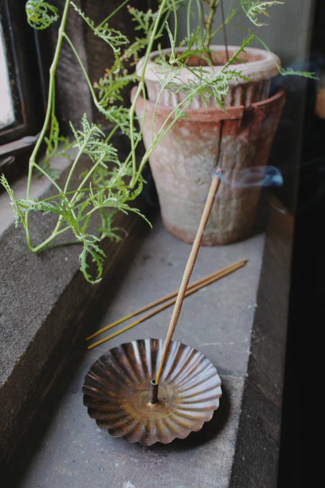 Scalloped metal incense holder with an aged finish, shown with a lit incense stick, incense sticks and terracotta pots.