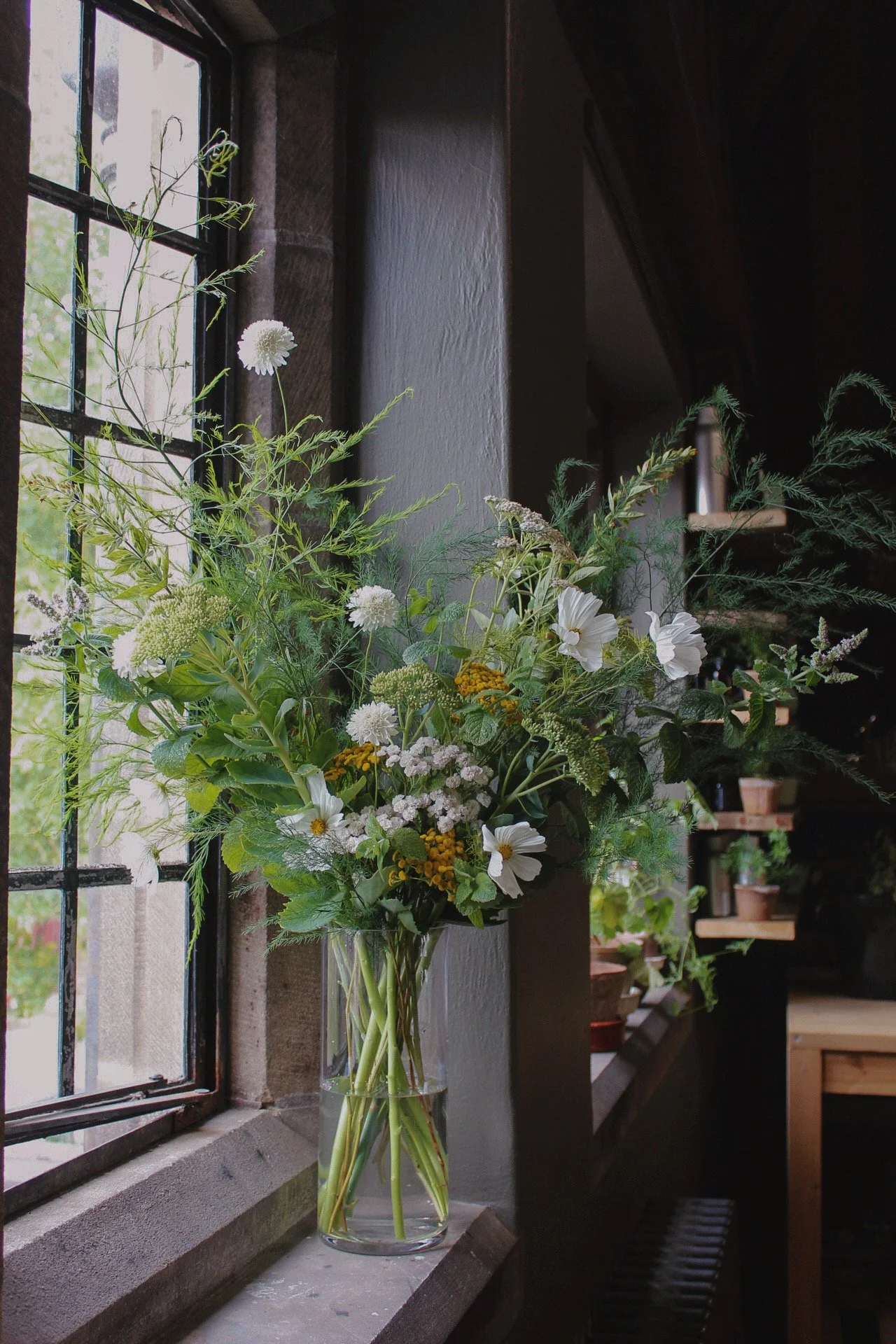 Tall glass grand centrepiece filled with seasonal white flowers, airy foliage and yellow meadow flowers beside a tall window.