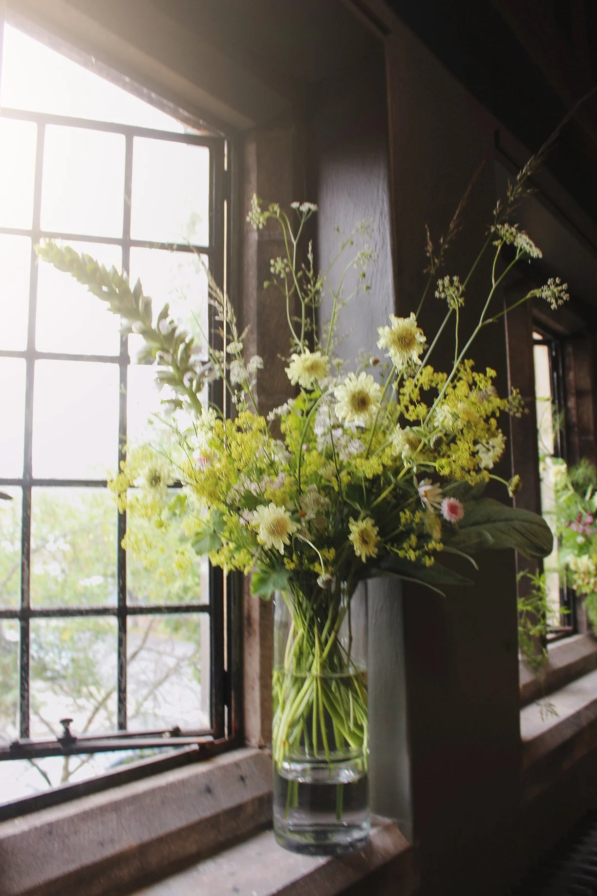 Oversized glass statement vase filled with abundant seasonal white and yellow flowers and foliage beside a tall window.