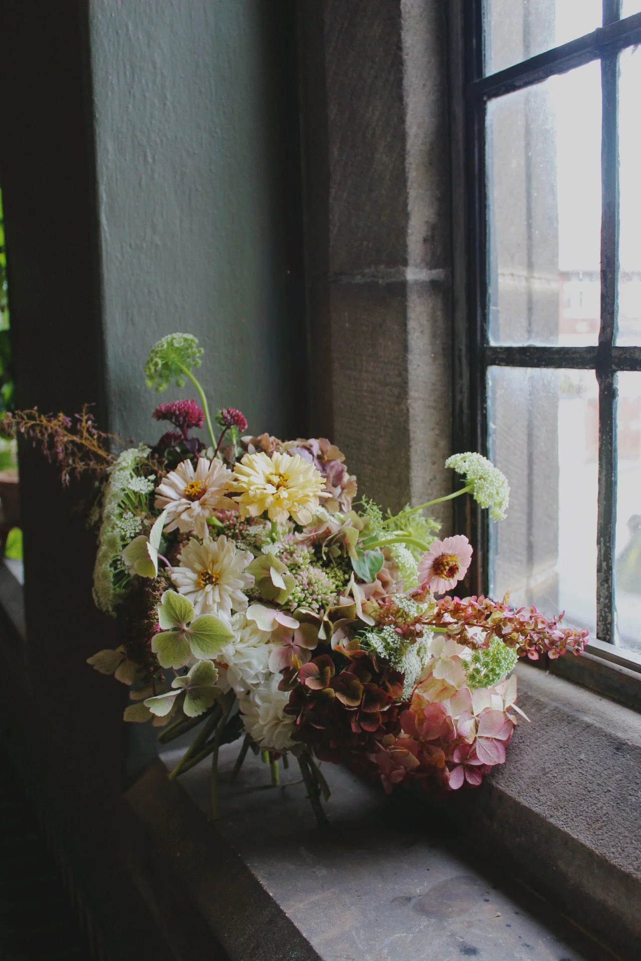A wedding bridal bouquet of flowers including hydrangeas, zinnias, and Queen Anne's lace placed on a windowsill next to a stone wall, with a black window frame and a rain-streaked window showing a blurry outdoor scene.