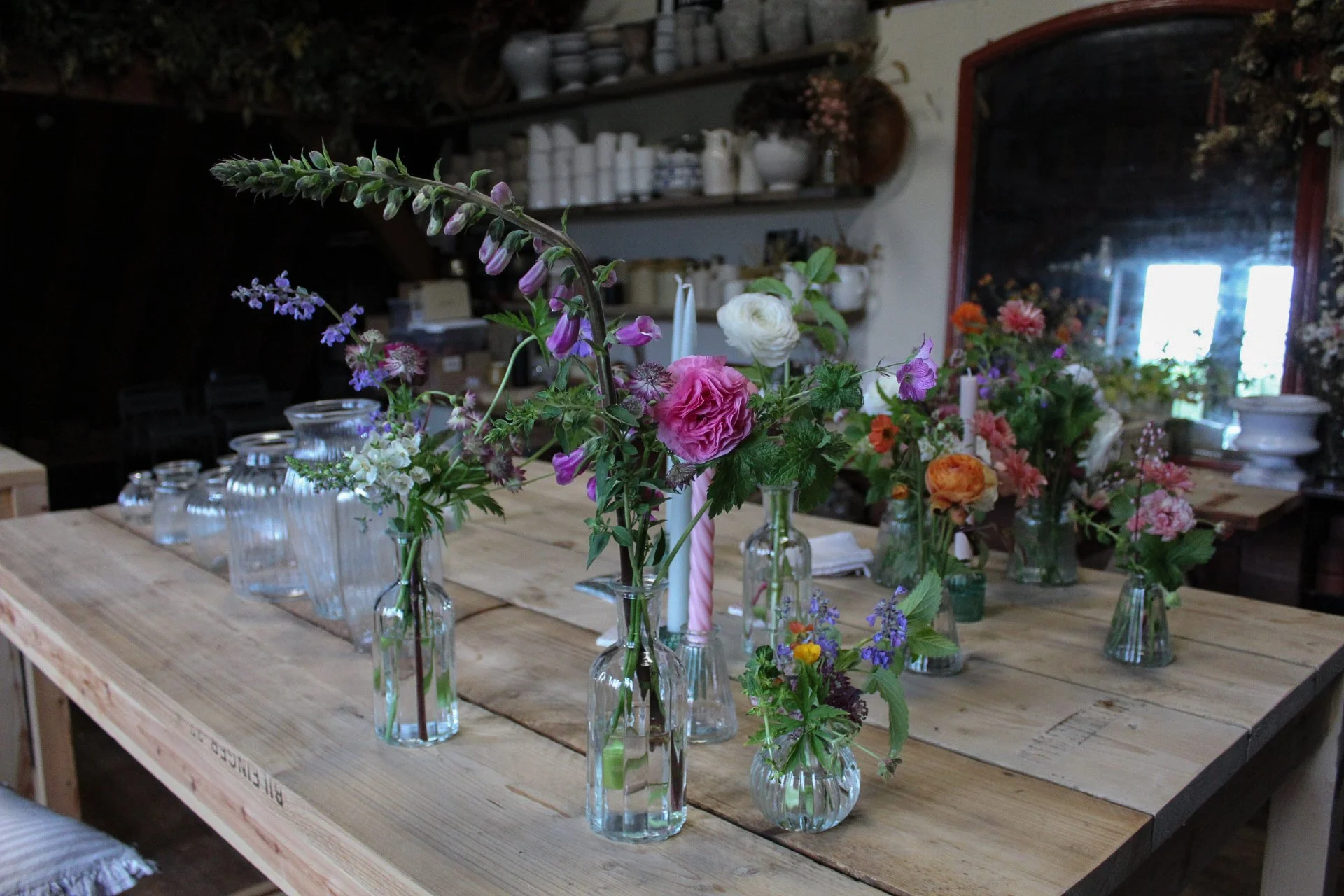 Various colorful wedding flowers arranged in glass vases on a wooden table in The Wild Potager co studio with shelves and a mirror in the background.